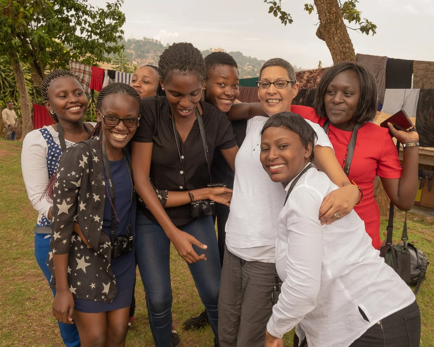 A group of women and girls smiling and hugging outdoors in a grassy area with trees and laundry drying on a line behind them.