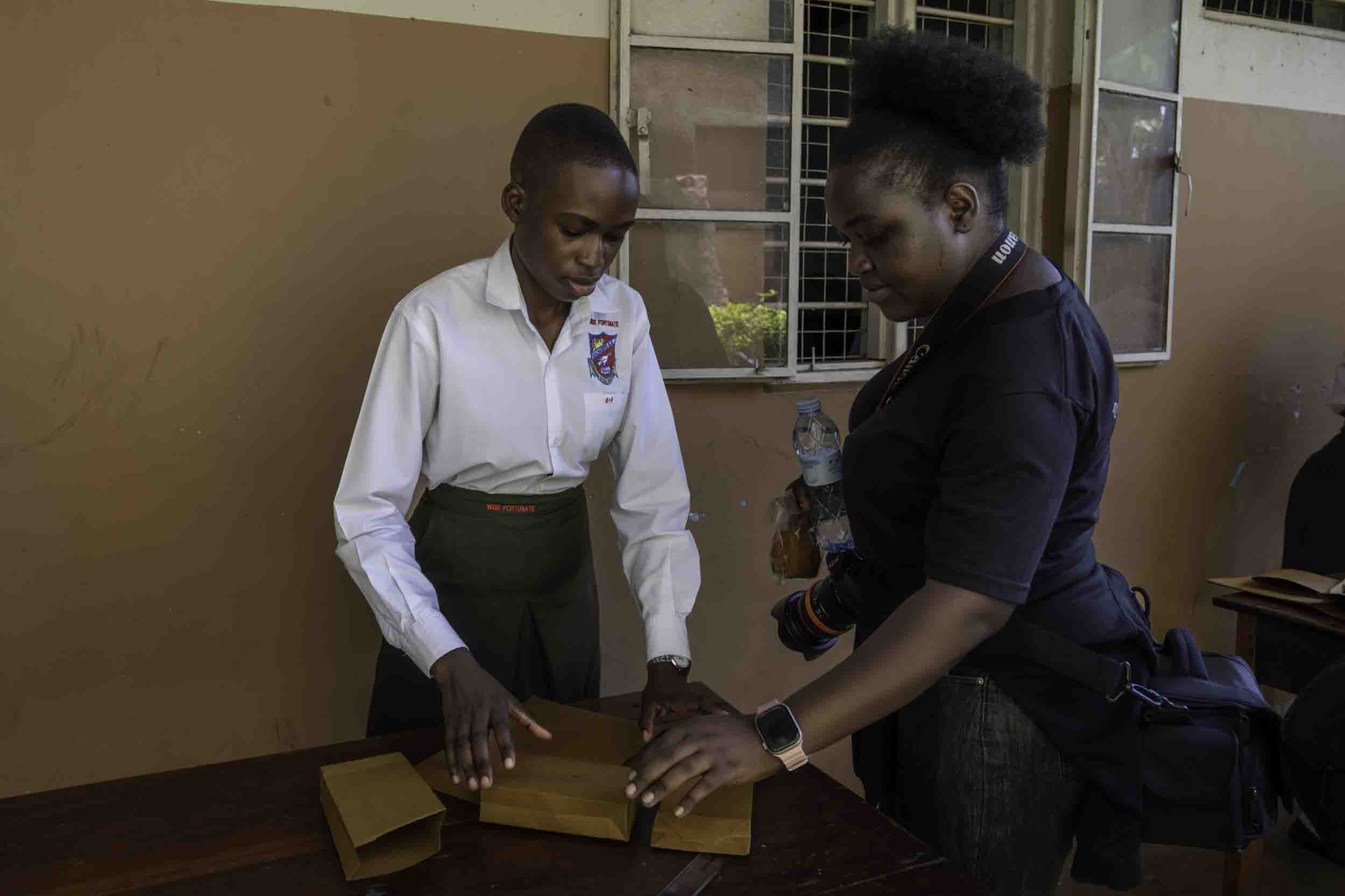 Two women are engaging in an activity with a wooden object in front of a beige wall with windows. One woman is wearing a white shirt and dark pants, and the other is wearing a dark shirt with a camera strap around her neck, holding a water bottle. Th