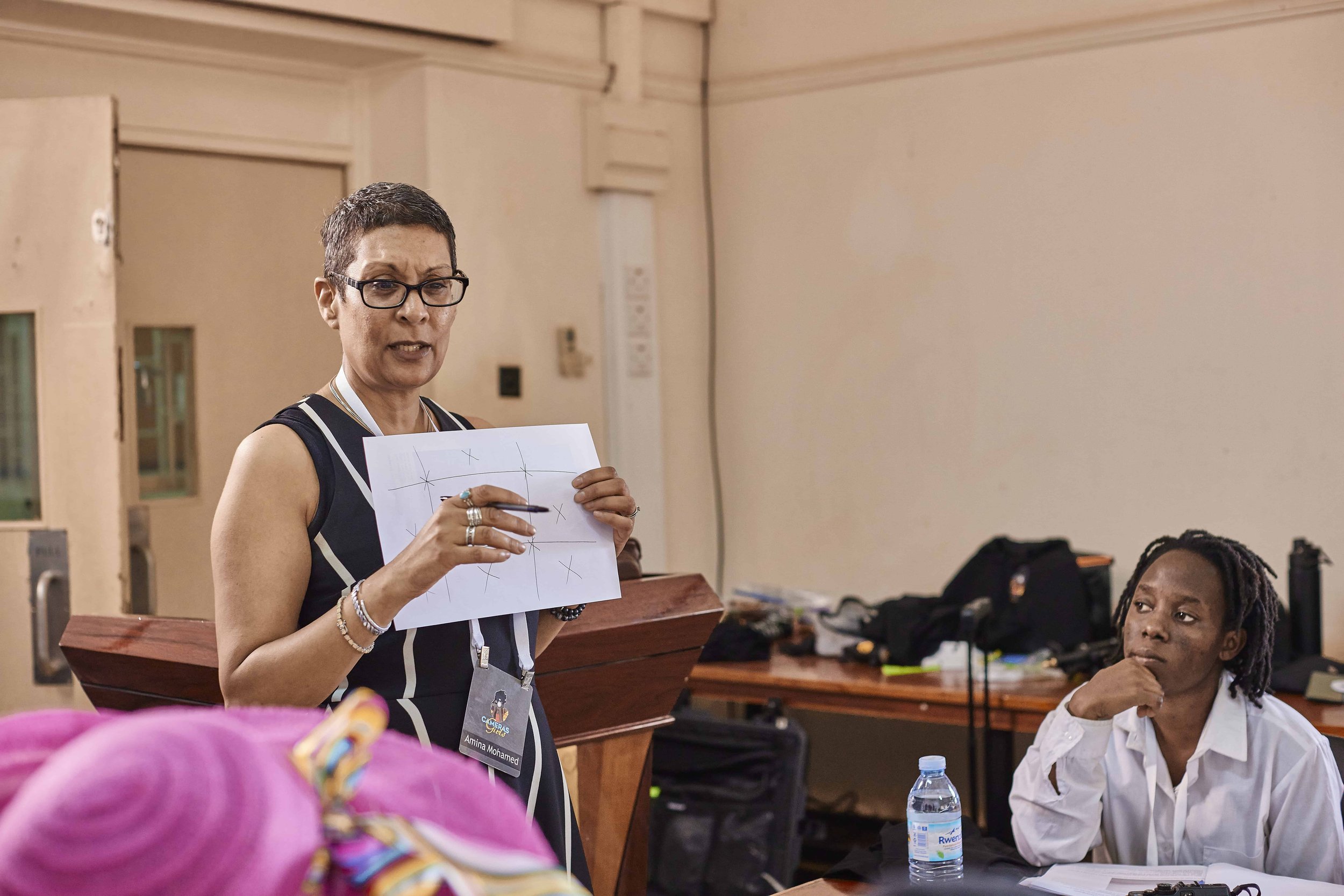 A woman with short hair, glasses, and a black and white vest is standing and holding a paper with a tic-tac-toe game written on it, in a room with beige walls and a wooden piano. Seated next to her is another woman with long dreadlocks, wearing a whi