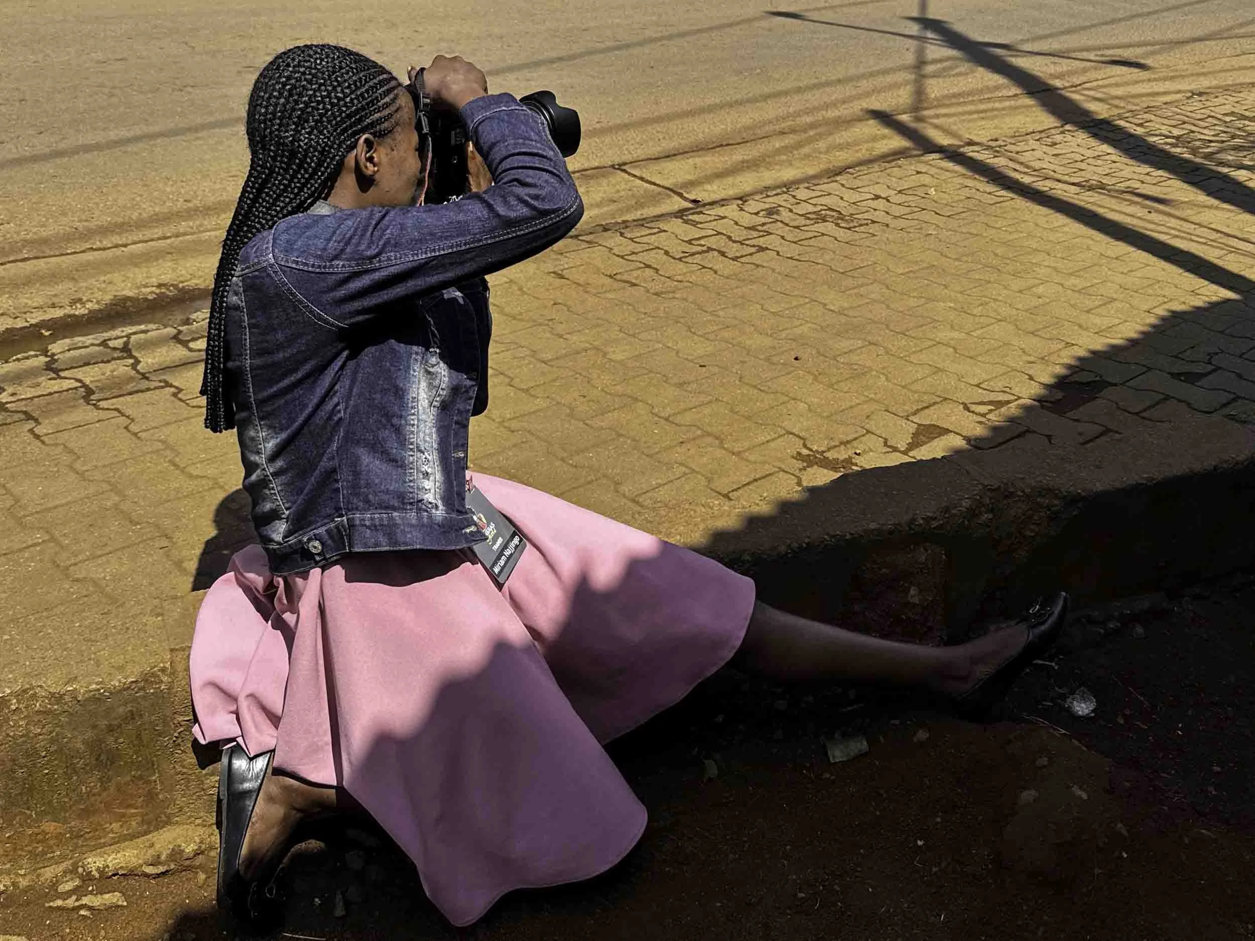 A woman with braided hair wearing a denim jacket and pink skirt, holding a camera and taking a photo outdoors on a brick sidewalk with some shadows.