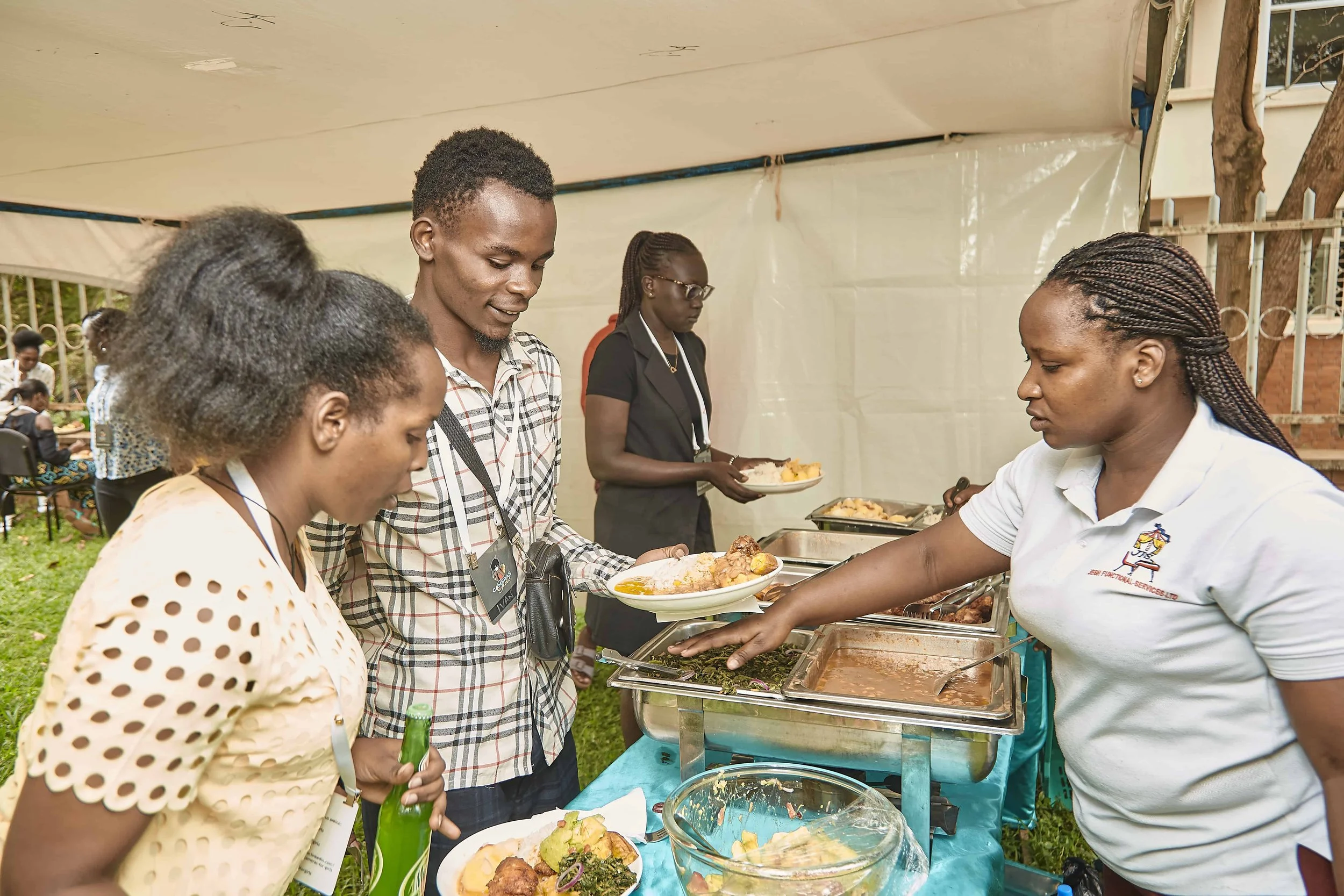 People standing in line at a buffet table outdoors, serving and receiving food at an event, with additional people in the background.