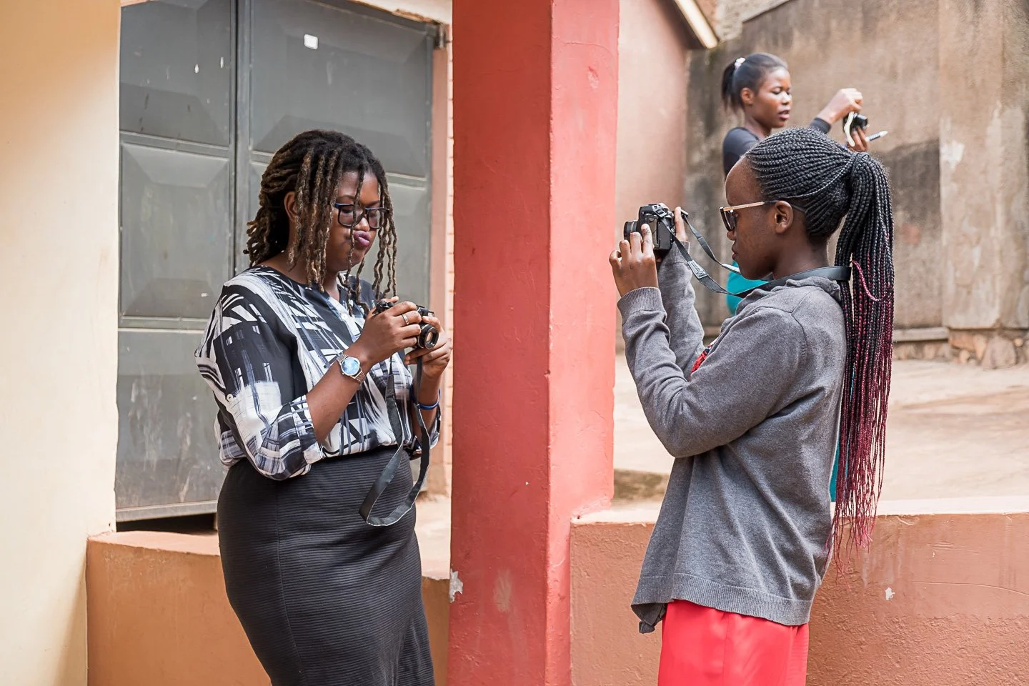 Three women are taking photos with cameras outdoors near a reddish wall and steps, in an urban setting.