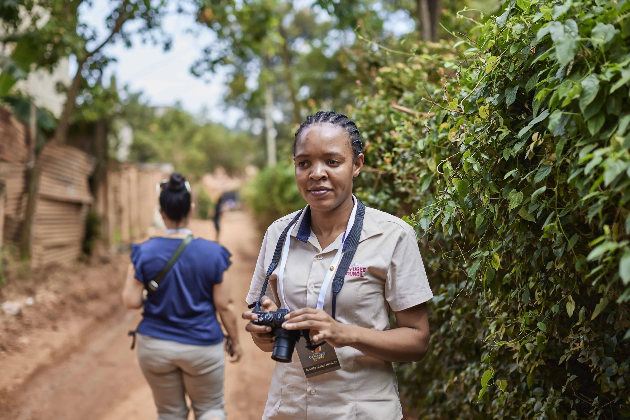 A woman with braided hair wearing a beige shirt with a 'Refugee Council' logo holding a camera, walking on a dirt path surrounded by greenery, with other people in the background.