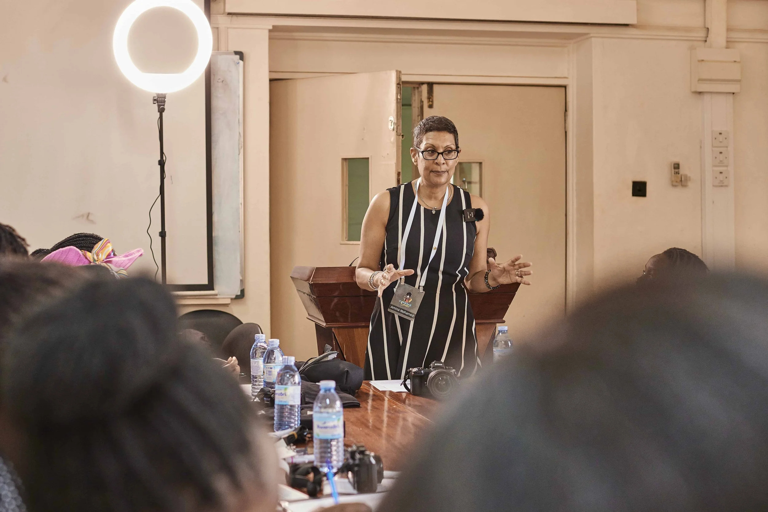 A woman with short hair, glasses, and a striped sleeveless dress is speaking or presenting in a room with a group of people. There are water bottles, a camera, and electronic devices on the table in front of her. A round light is visible on a stand t