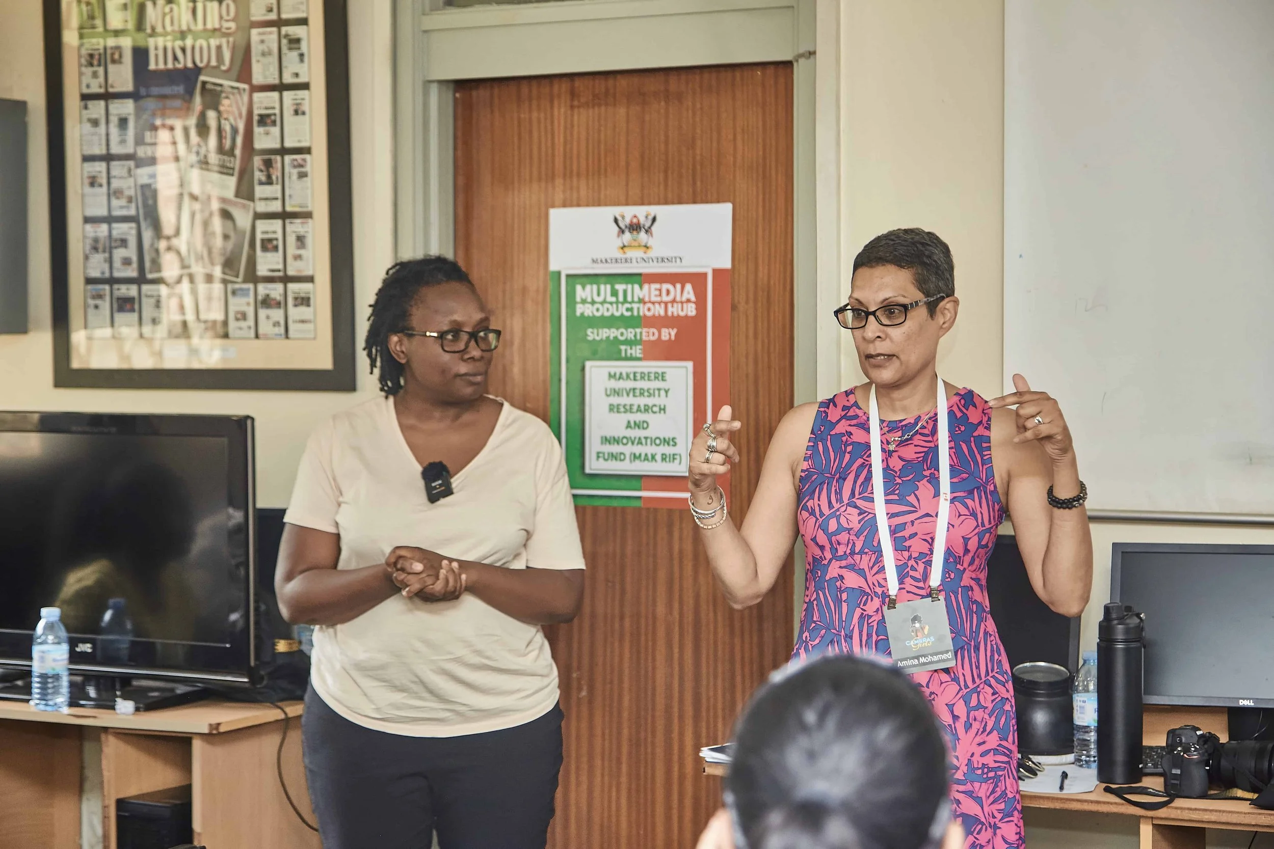 Two women are engaged in a discussion or presentation in a classroom setting. One woman is gesturing with her hands, while the other woman is listening. There are posters and computer monitors visible in the background.