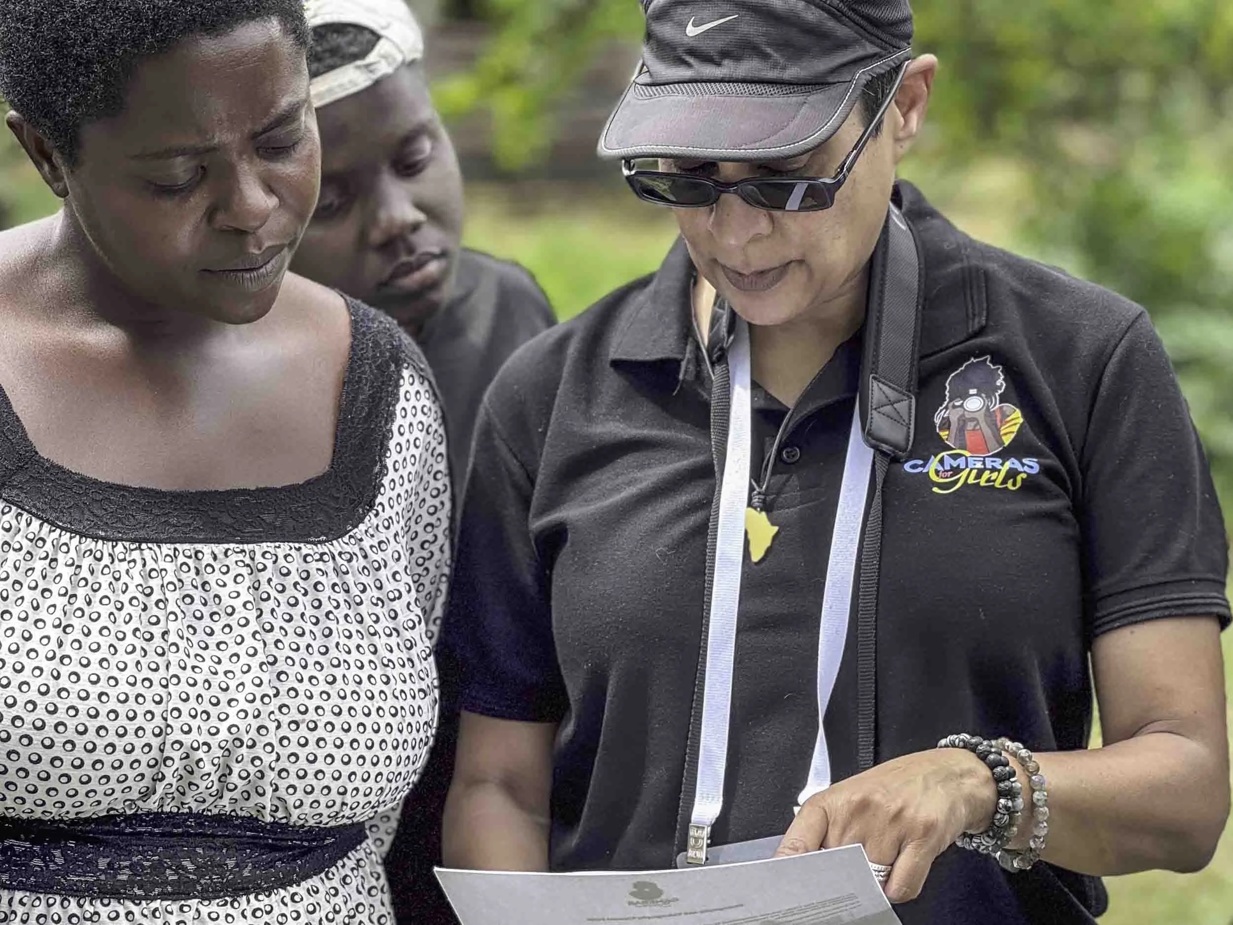 Three women closely looking at a paper or photograph, with one wearing sunglasses, a black cap, and a black shirt with a logo. The scene appears to be outdoors with greenery in the background.