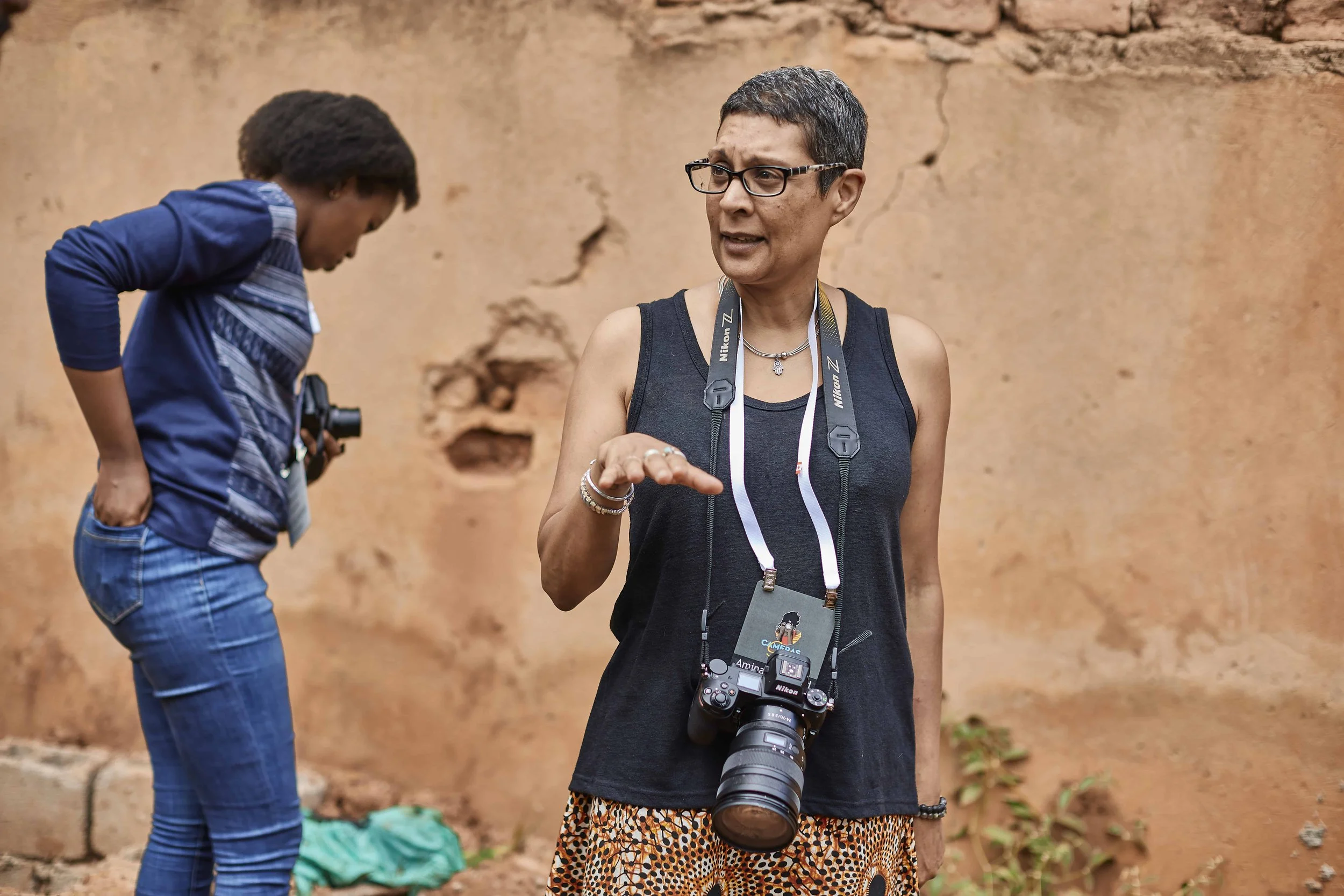 Two women, one with glasses and camera around neck, speaking while another is looking down and handling something in her hands, against a mud wall.