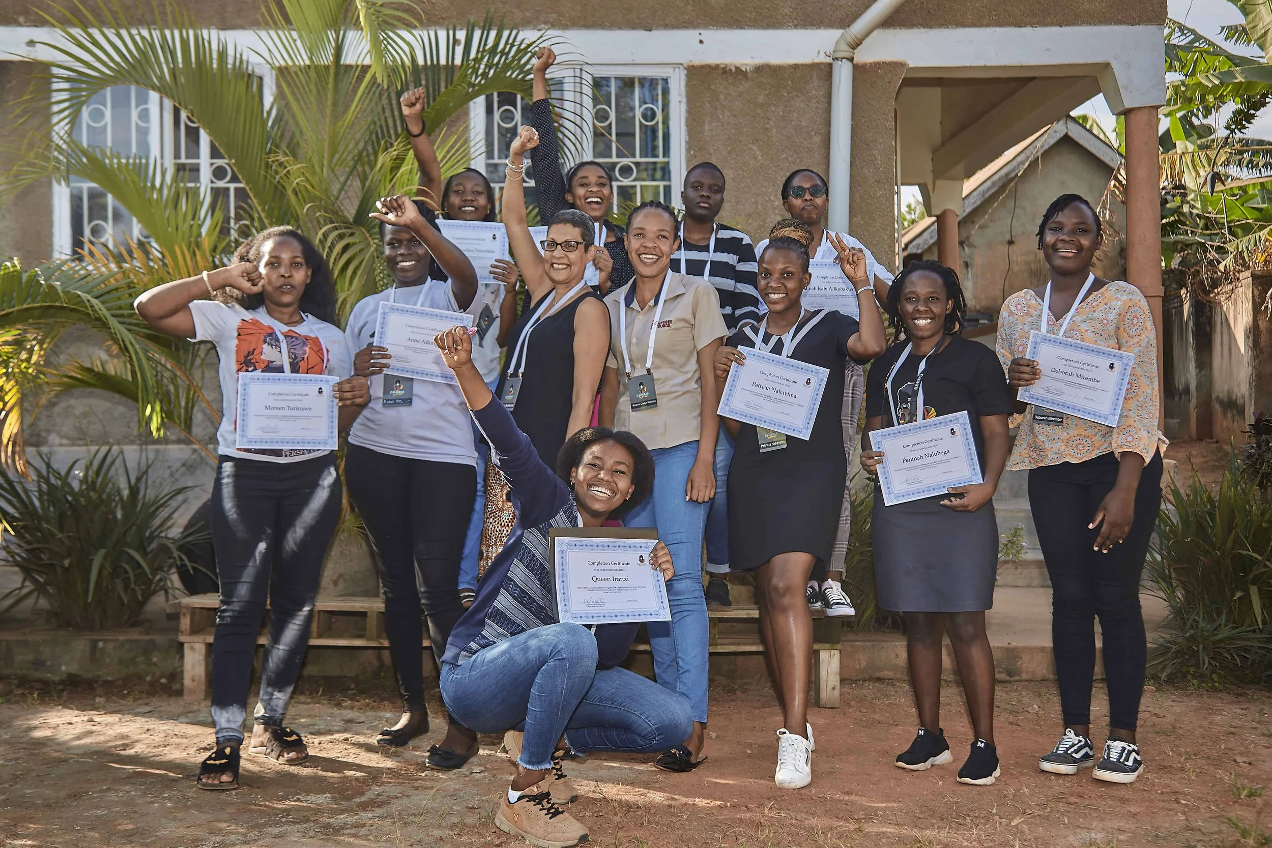 Group of women posing outdoors, many holding certificates and smiling, some raising fists, with house and plants in the background.