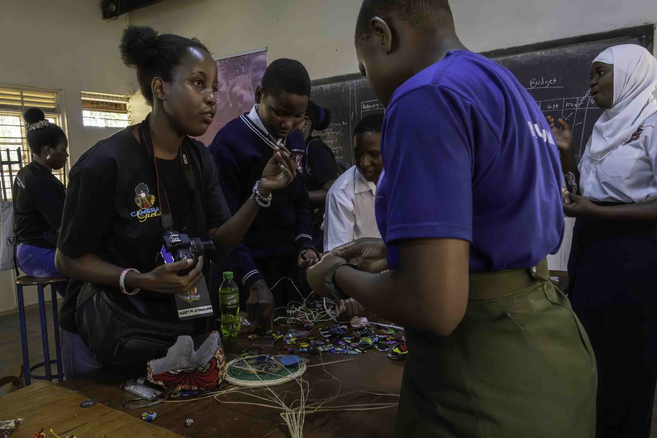 Group of students and a teacher in a classroom, with tables displaying handmade jewelry and crafts, engaged in a hands-on activity.