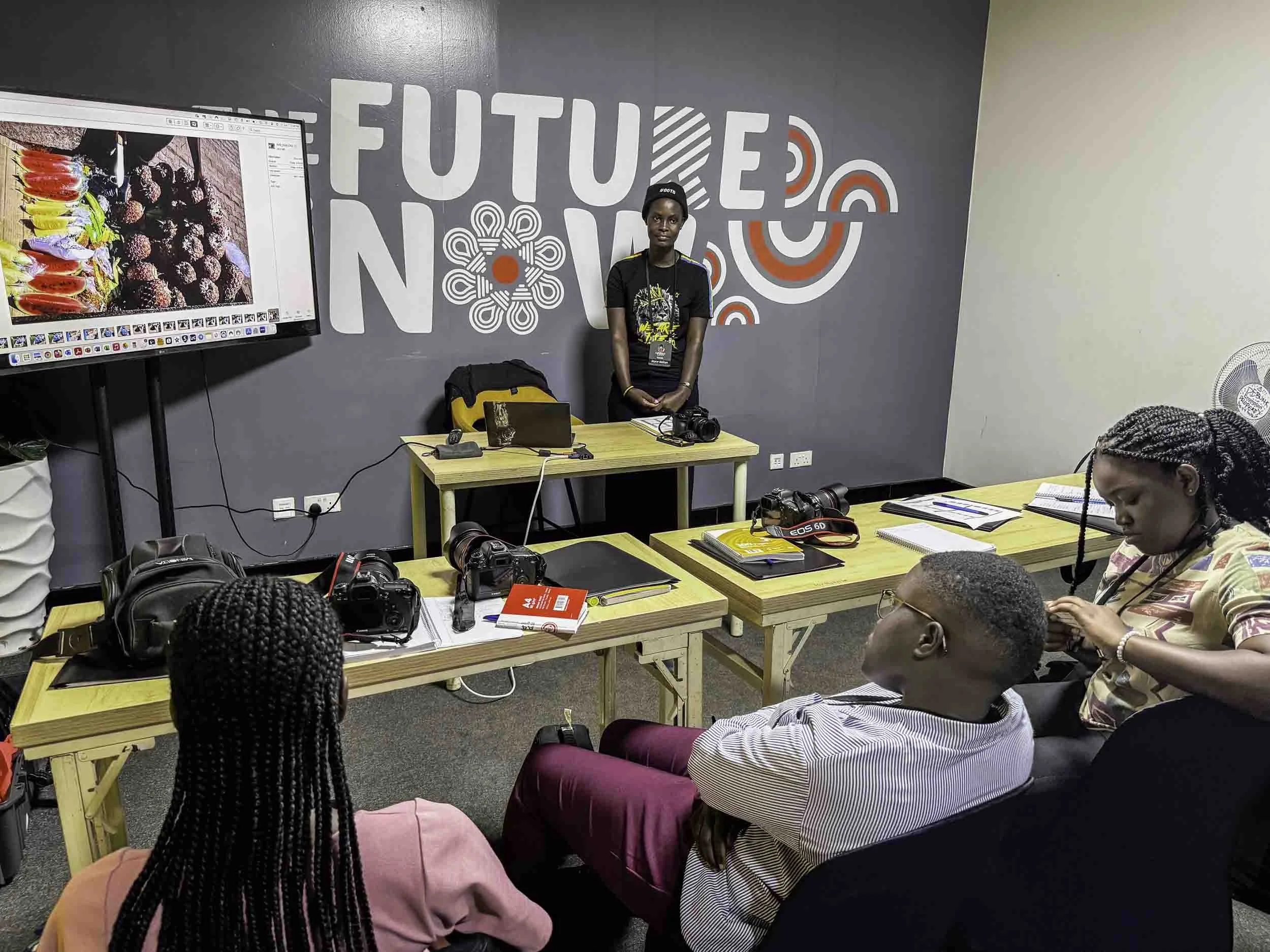 A woman standing in front of a wall that says 'Future Now' with abstract red and white designs. She is surrounded by seated people, some with cameras and notebooks on a table, in a classroom or conference room setting. There is a large screen display