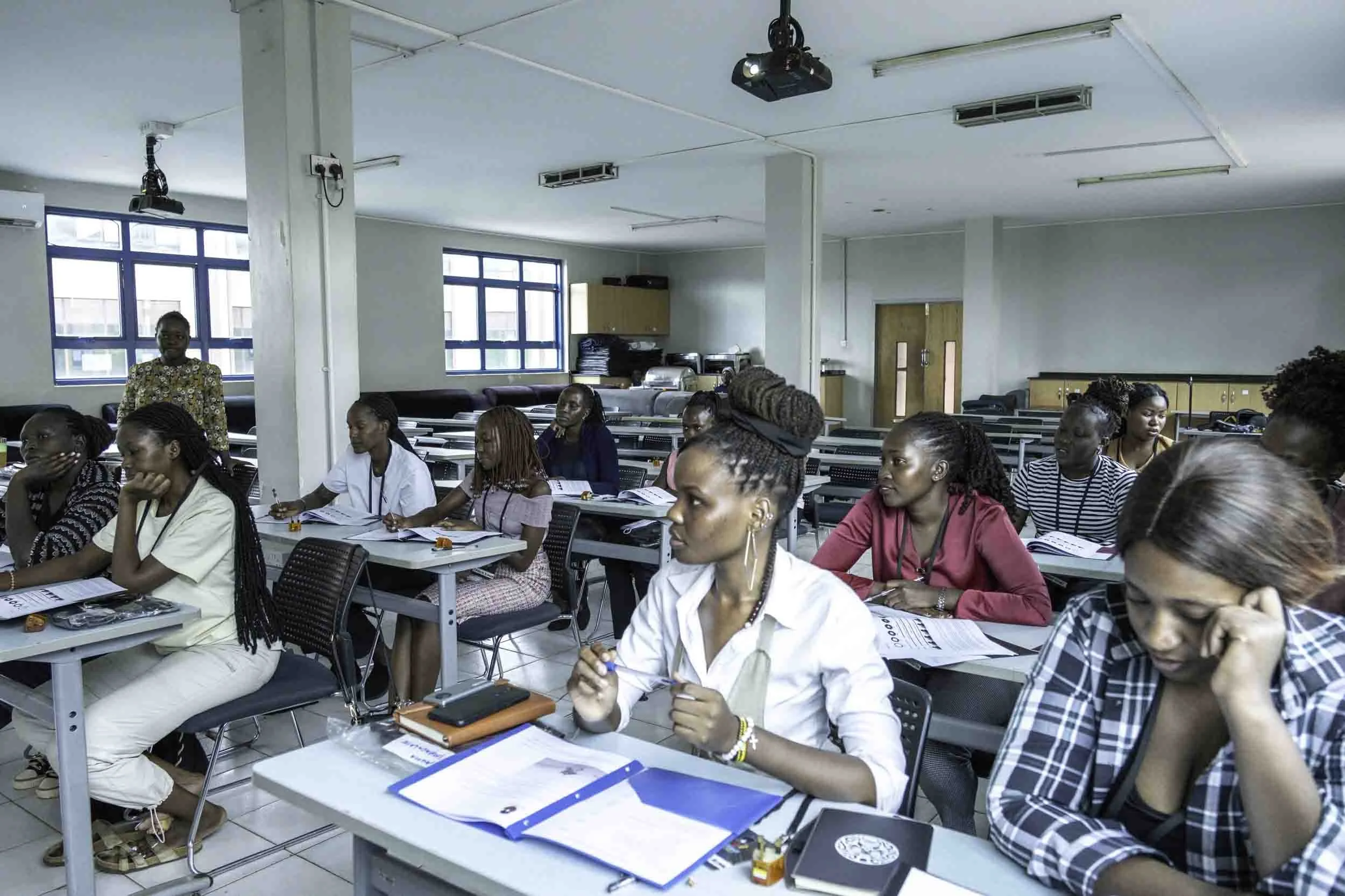 A classroom filled with mostly women students seated at desks, some taking notes or looking forward. There is a woman standing at the front, possibly the instructor, and windows behind her.