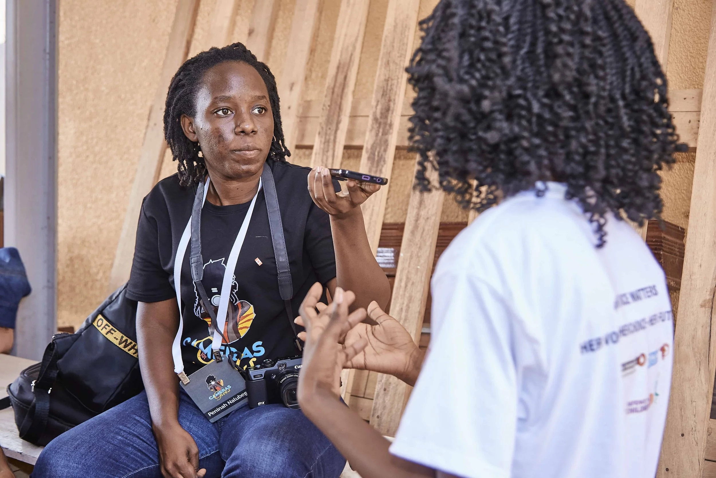 A woman sitting and talking to a person with curly hair, using a smartphone to record or interview her, in a setting with wooden walls.