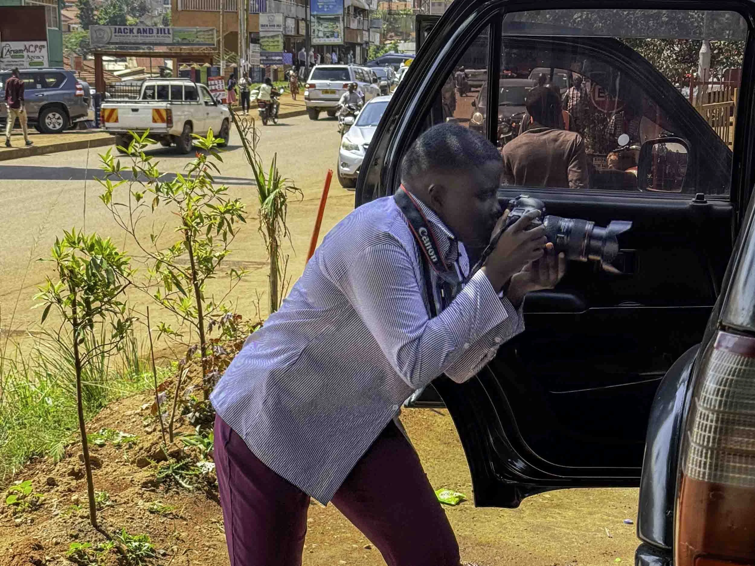 A man leaning into a vehicle, using a camera with a long lens. The scene appears to be in a busy outdoor area with several cars, people walking, and shops in the background.