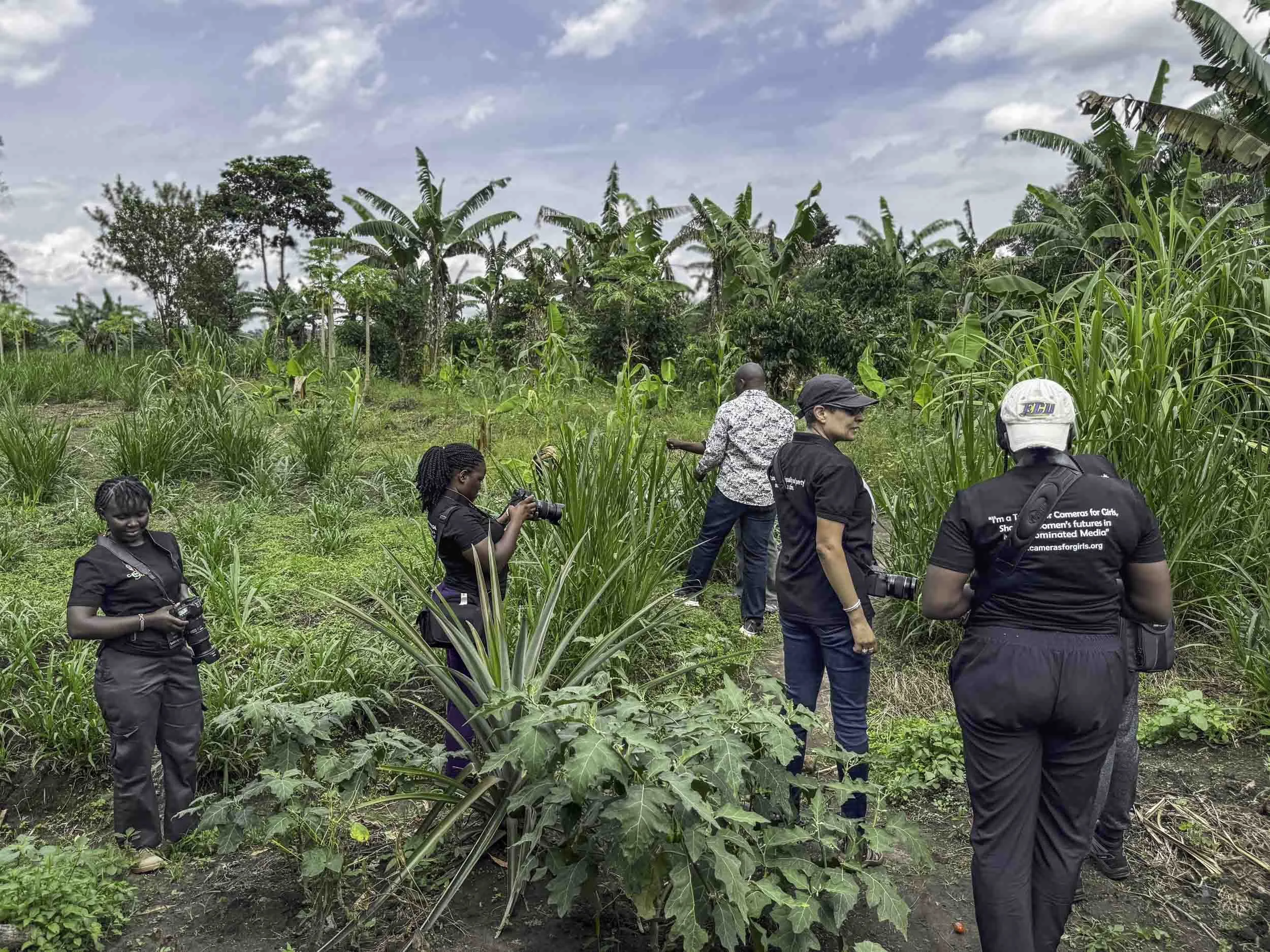 A group of people, including women and children, walking among banana and tall grass plants in a lush green outdoor setting under a partly cloudy sky.