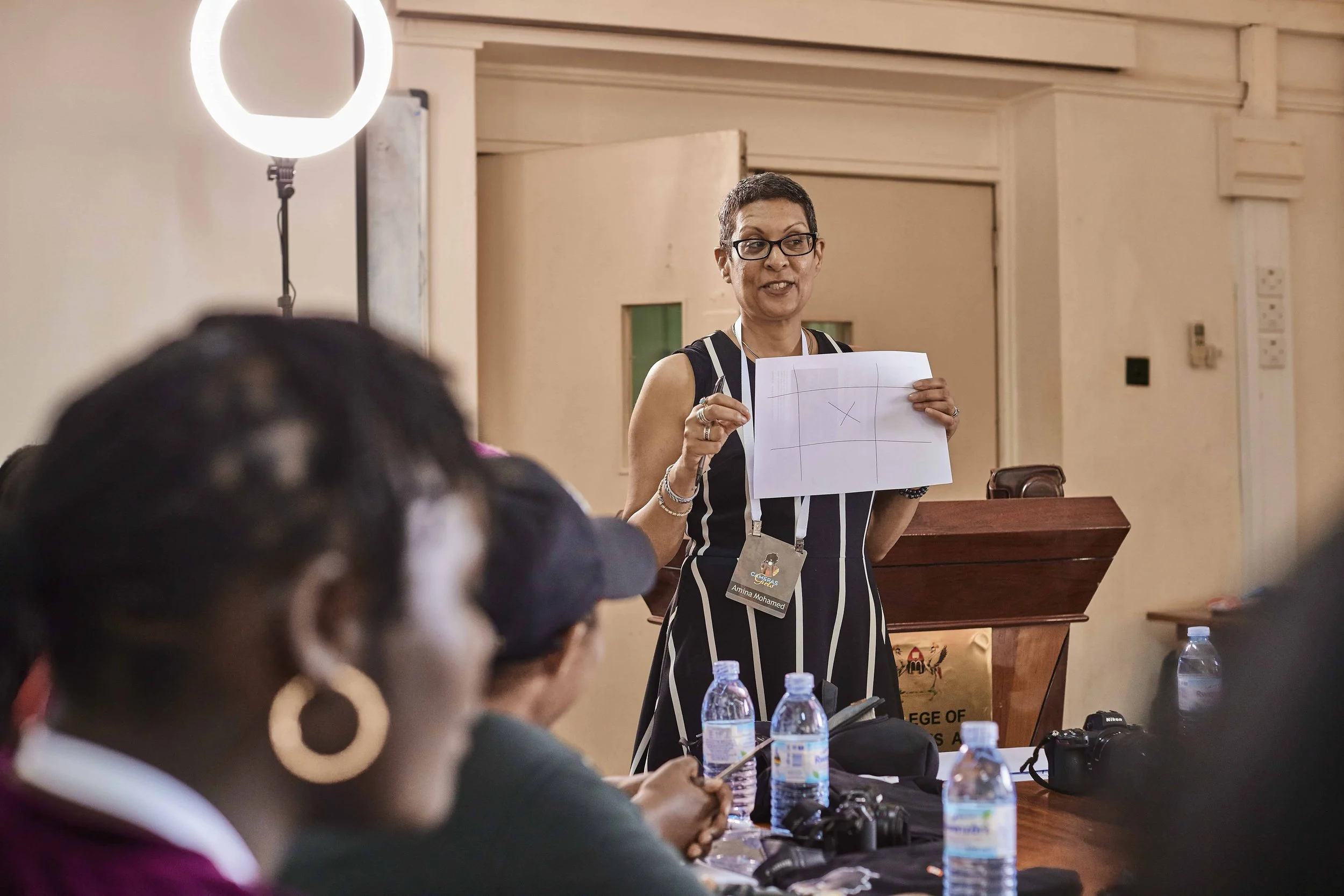 A woman wearing glasses and a striped dress is giving a presentation with a paper in her hand, while facing a seated audience in a conference room. There are water bottles and cameras on the table.