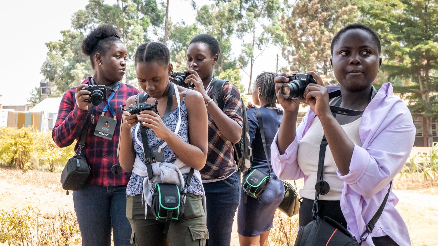 Five young women stand outdoors, each holding a camera and looking at their photos, with trees and a building in the background.