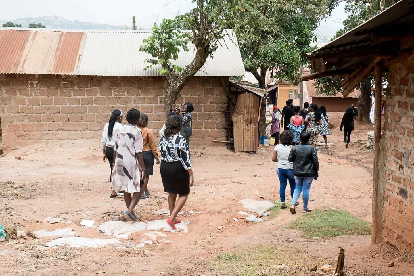 Group of women walking through a rustic village with brick and mud buildings, some trees, and an unpaved dirt ground.