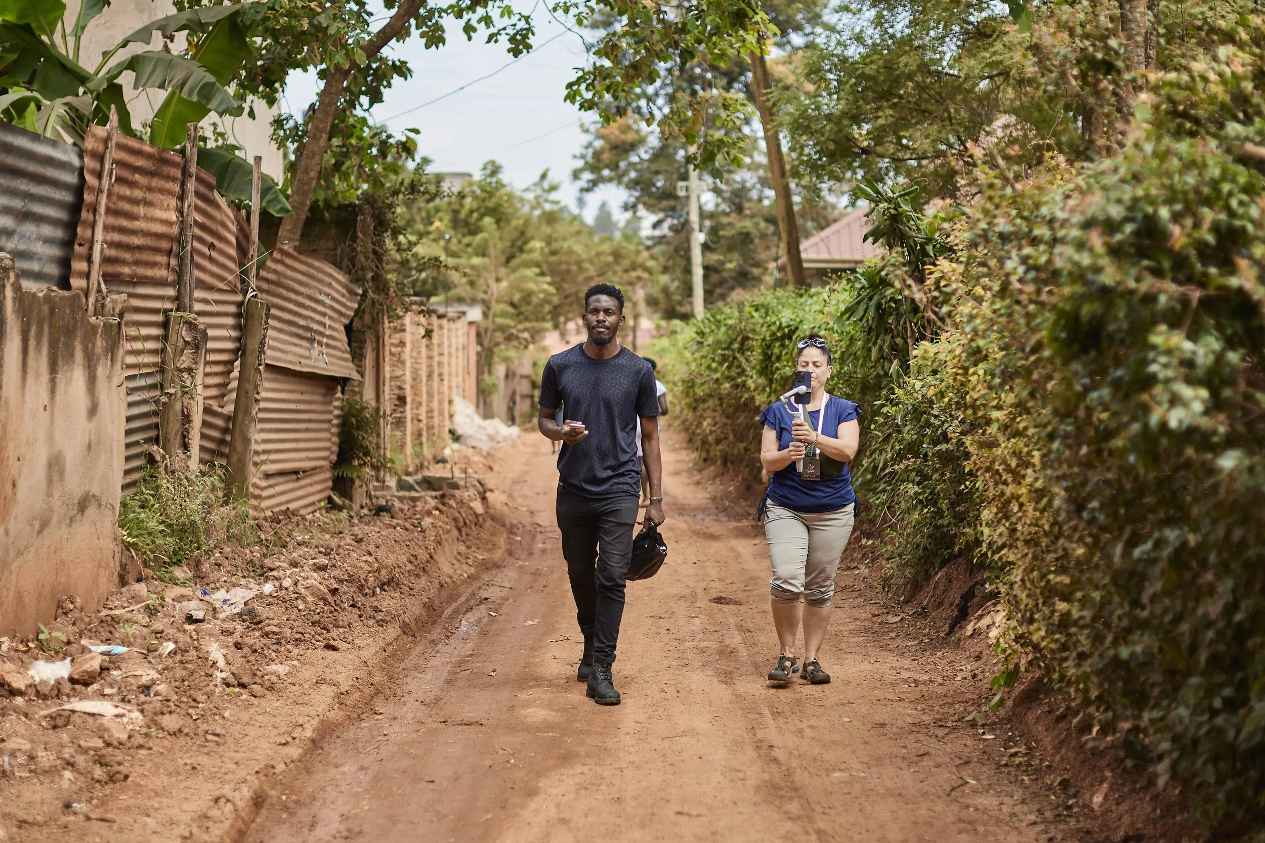 Two people walking along a dirt path in a rural area, with trees and bushes on either side. One person is holding a phone, and the other is holding a small bag, and they appear to be engaged in conversation.