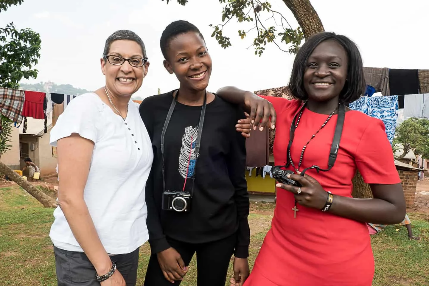 Three women standing outdoors, smiling for the camera, with laundry hanging in the background and trees around them.