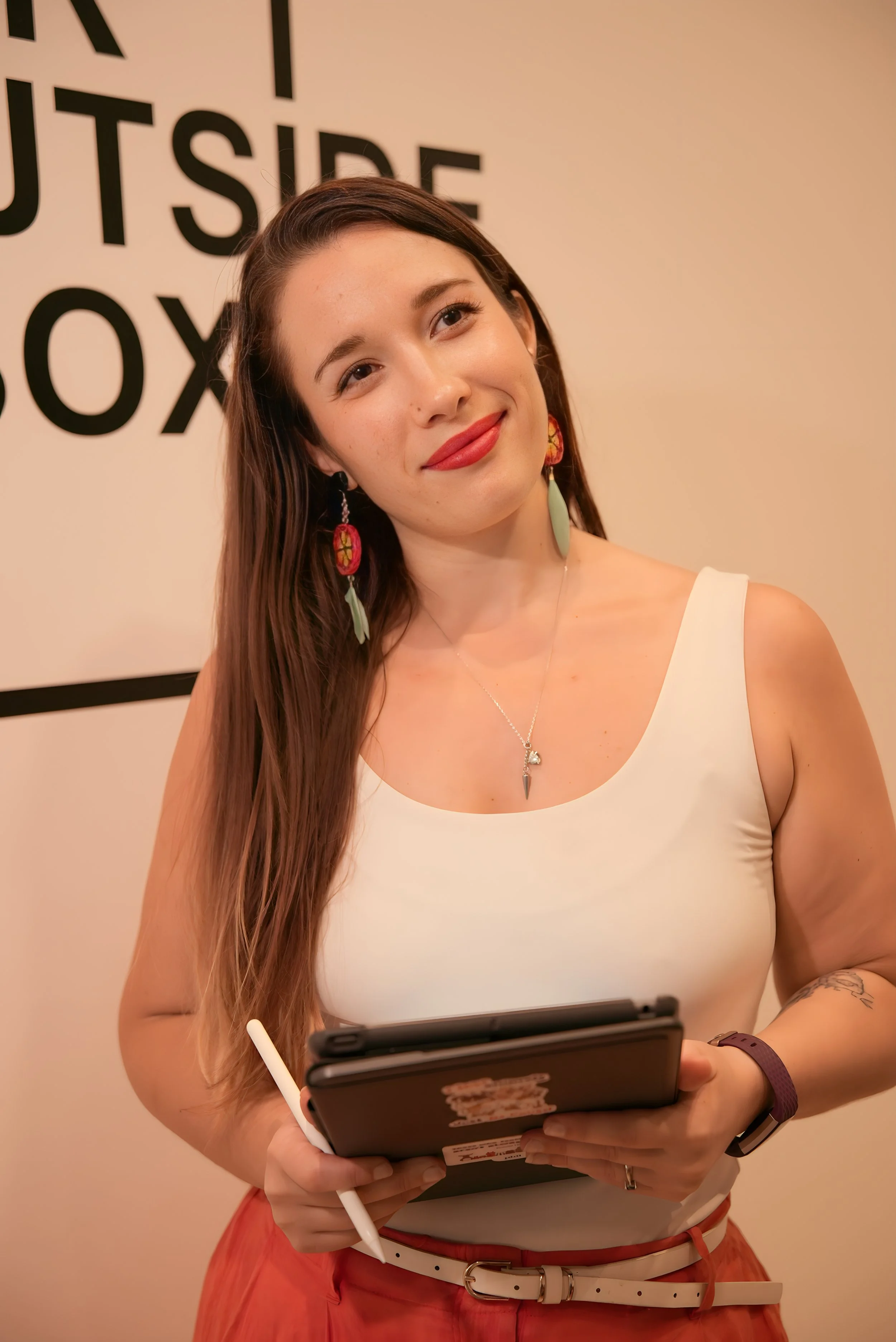 A young woman with long brown hair, wearing a white top and orange skirt with a white belt, is looking at a tablet. She has earrings, a necklace, a watch, and is holding a stylus. Behind her, there is a beige wall with text that says "INSIDE".