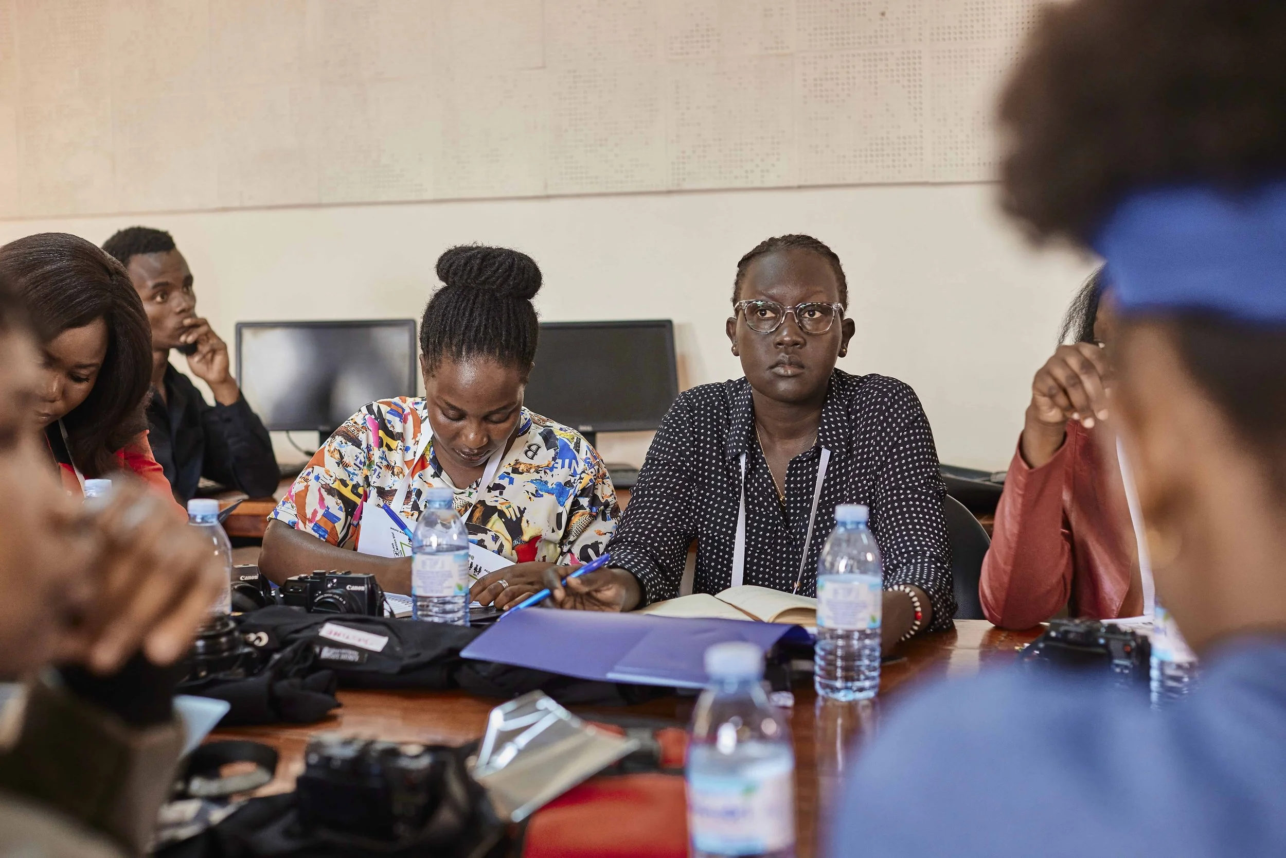 A group of diverse women sitting around a table engaged in a meeting or discussion, with notebooks, water bottles, and cameras on the table.