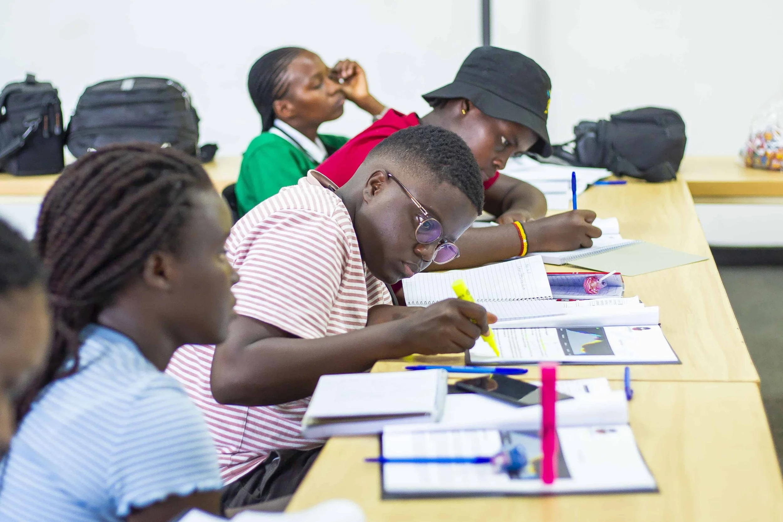 Students sitting at a table in a classroom, engaging with notebooks and colorful pens, with backpacks placed on the table.
