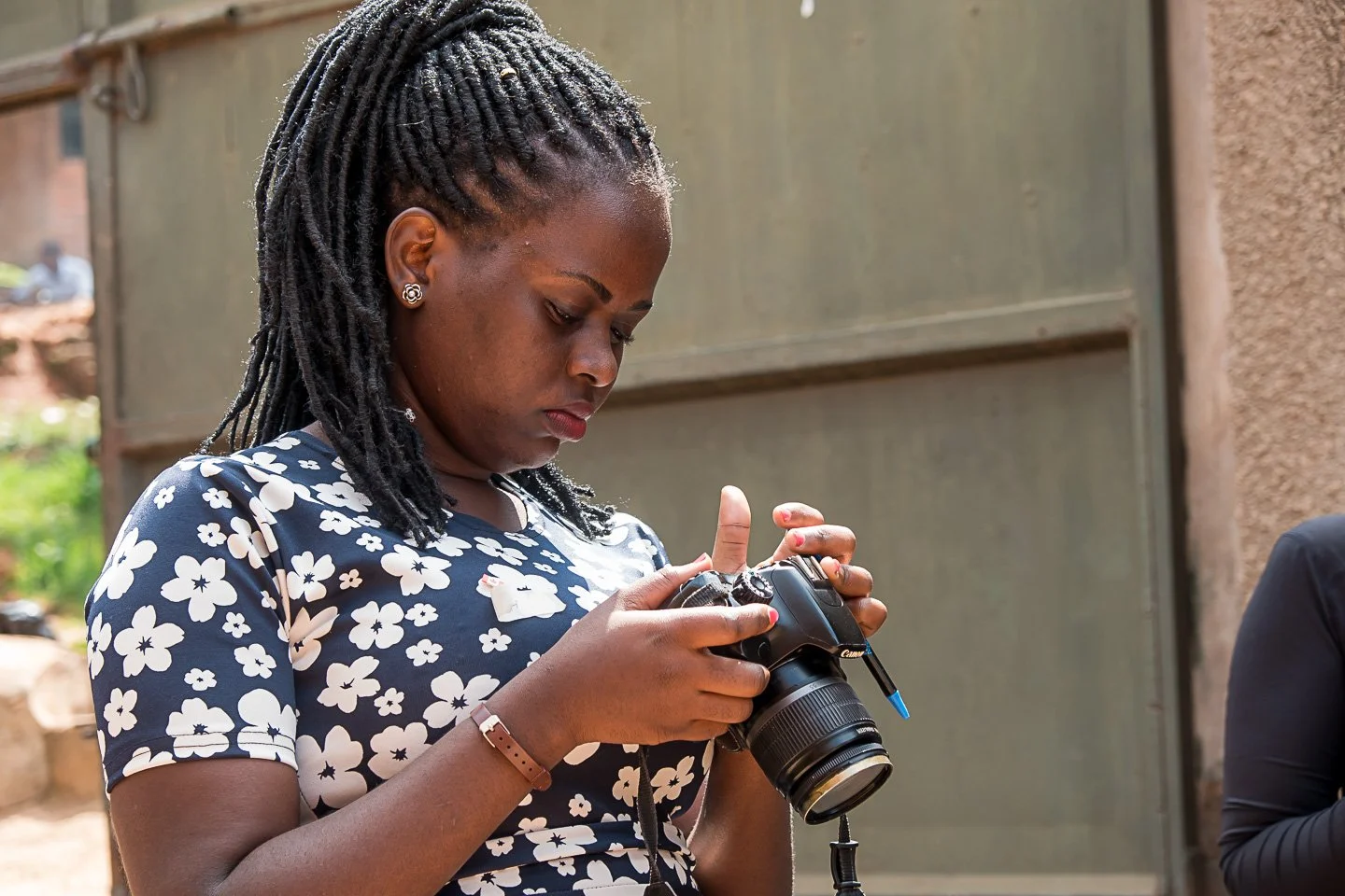 A woman with dreadlocks wearing a navy blue dress with white floral pattern, looking at a DSLR camera she is holding in her hands.