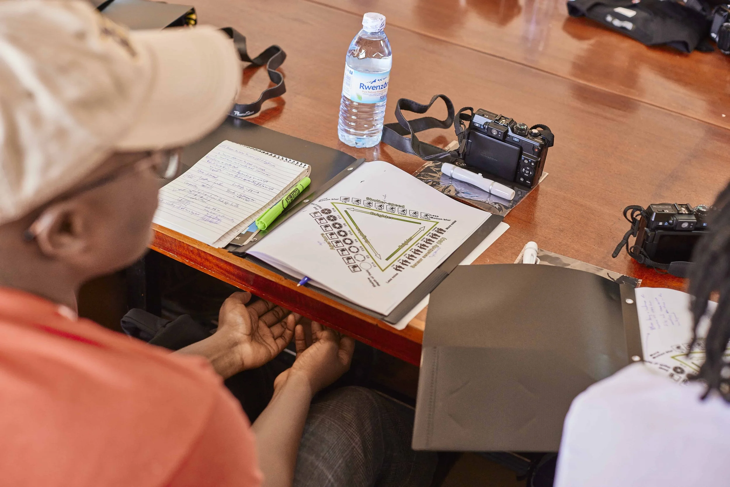 A person with glasses and light-colored hair is sitting at a wooden table, holding another person's hand. On the table are notebooks, highlighters, a water bottle, a camera, and some papers. The background includes more tables and supplies.