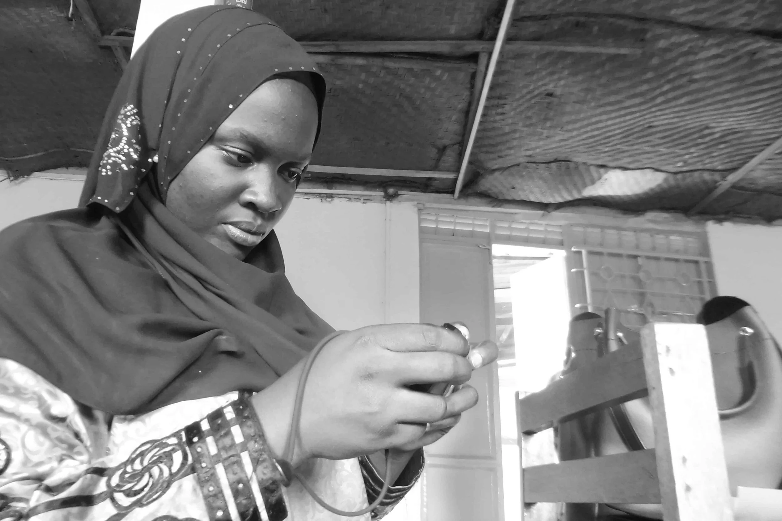 A woman wearing a headscarf is working with a sewing machine indoors, with a wooden ceiling and some windows in the background.
