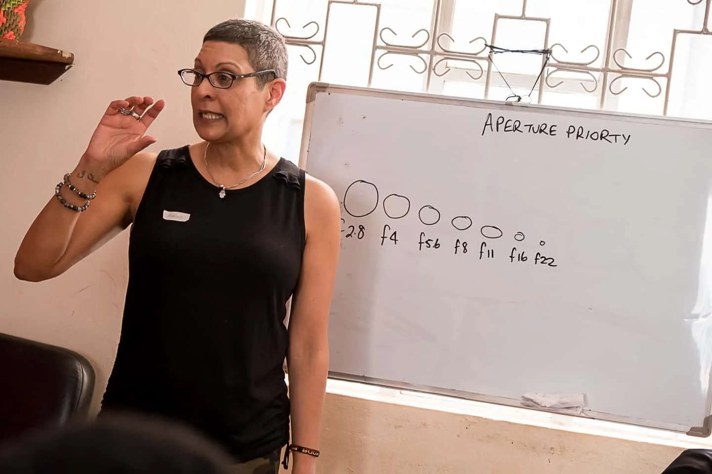 A woman standing in front of a whiteboard with the title 'Aperture Priority' and various aperture sizes written below it. She appears to be giving a presentation or lecture.