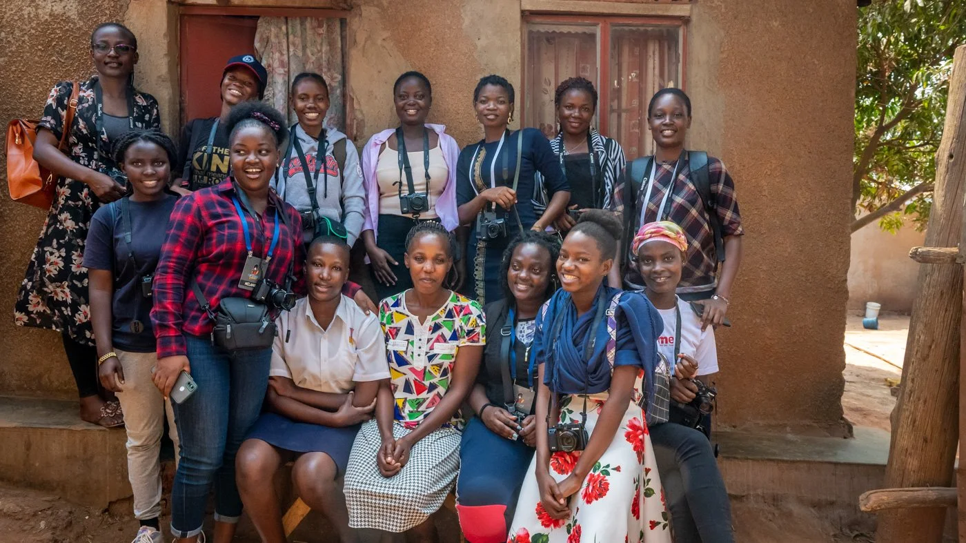 A group of women and girls standing and sitting outside a mud-brick building, some holding cameras and wearing backpacks.
