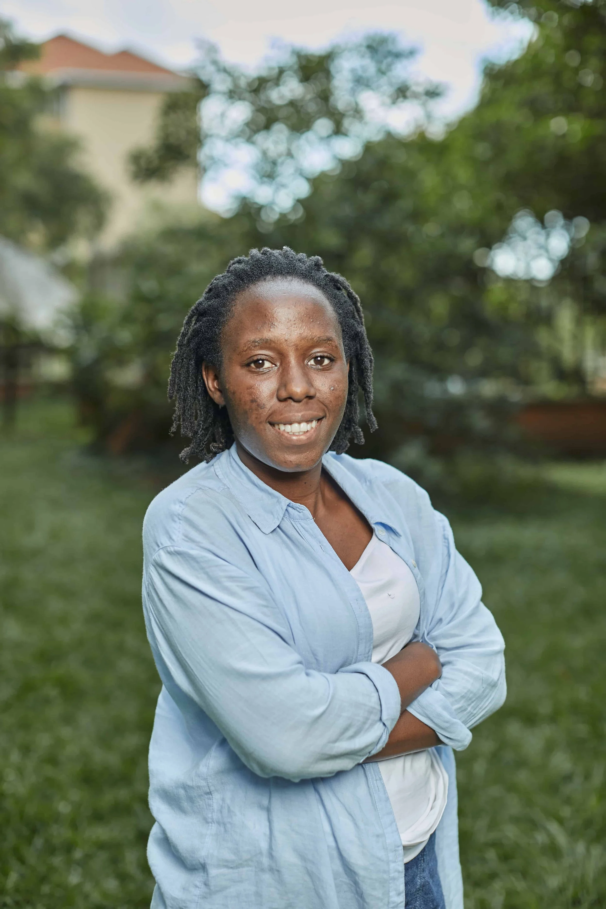 A woman with dreadlocks wearing a light blue shirt standing outdoors with arms crossed, smiling, with greenery and a house in the background.