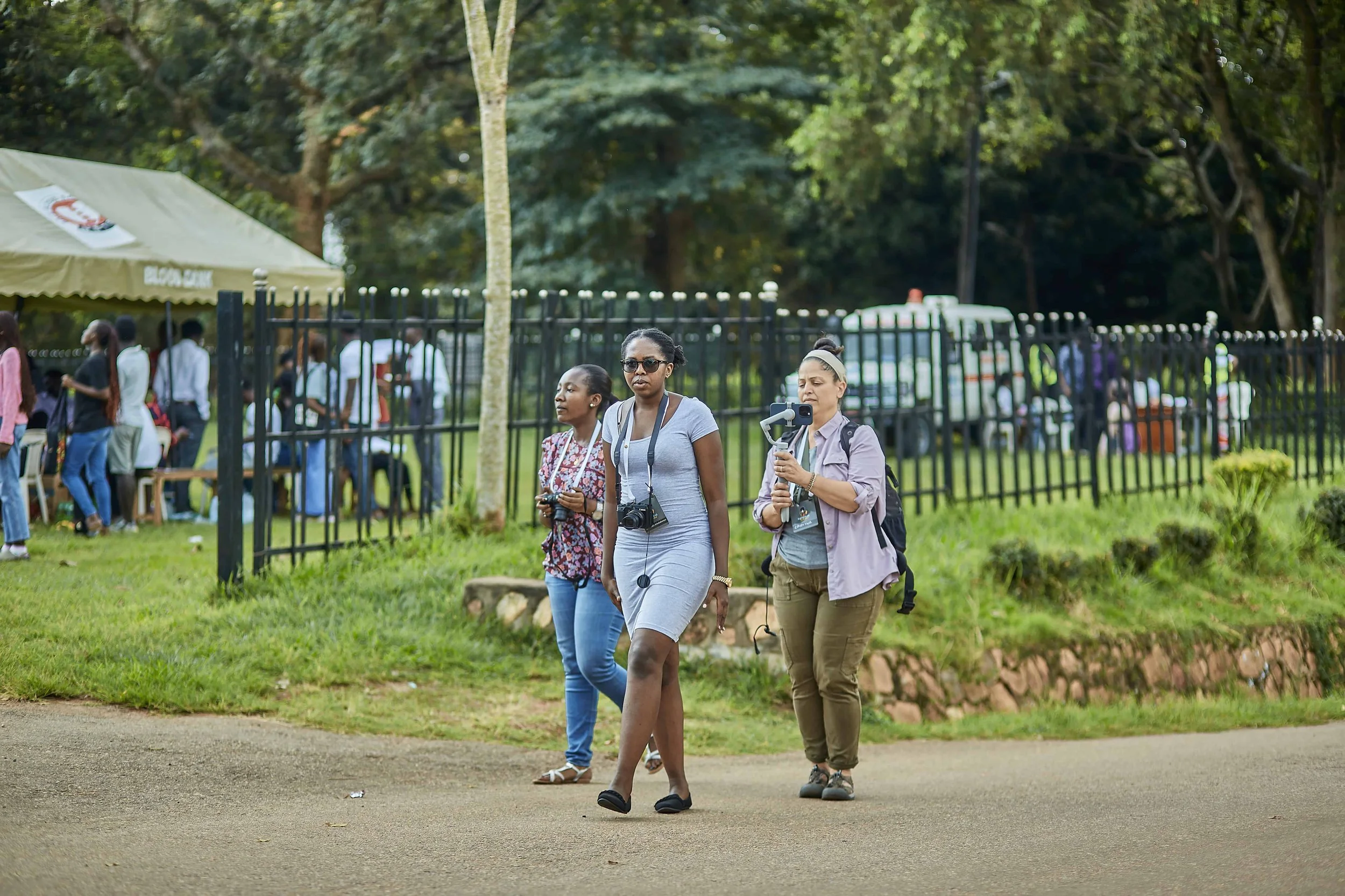 Three women walking on a park pathway with trees and a black fence in the background; one woman is taking a photo or recording a video with a handheld device.