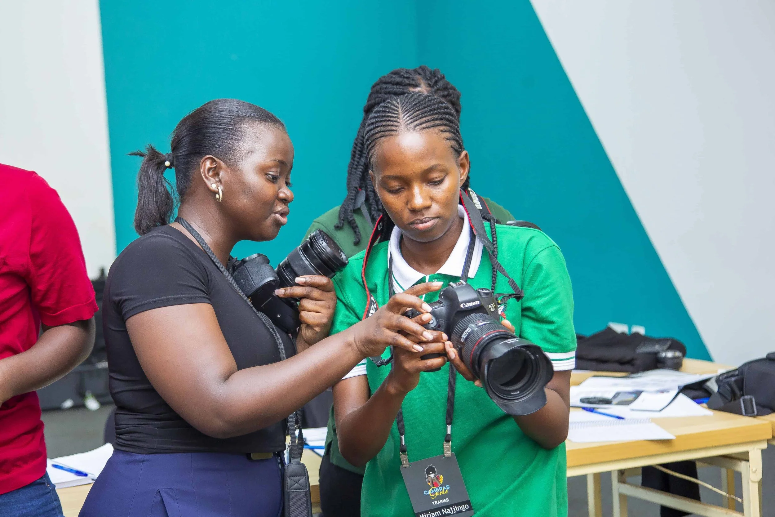 Two women review a professional camera, their heads close together, at an indoor event with teal walls and tables with equipment.