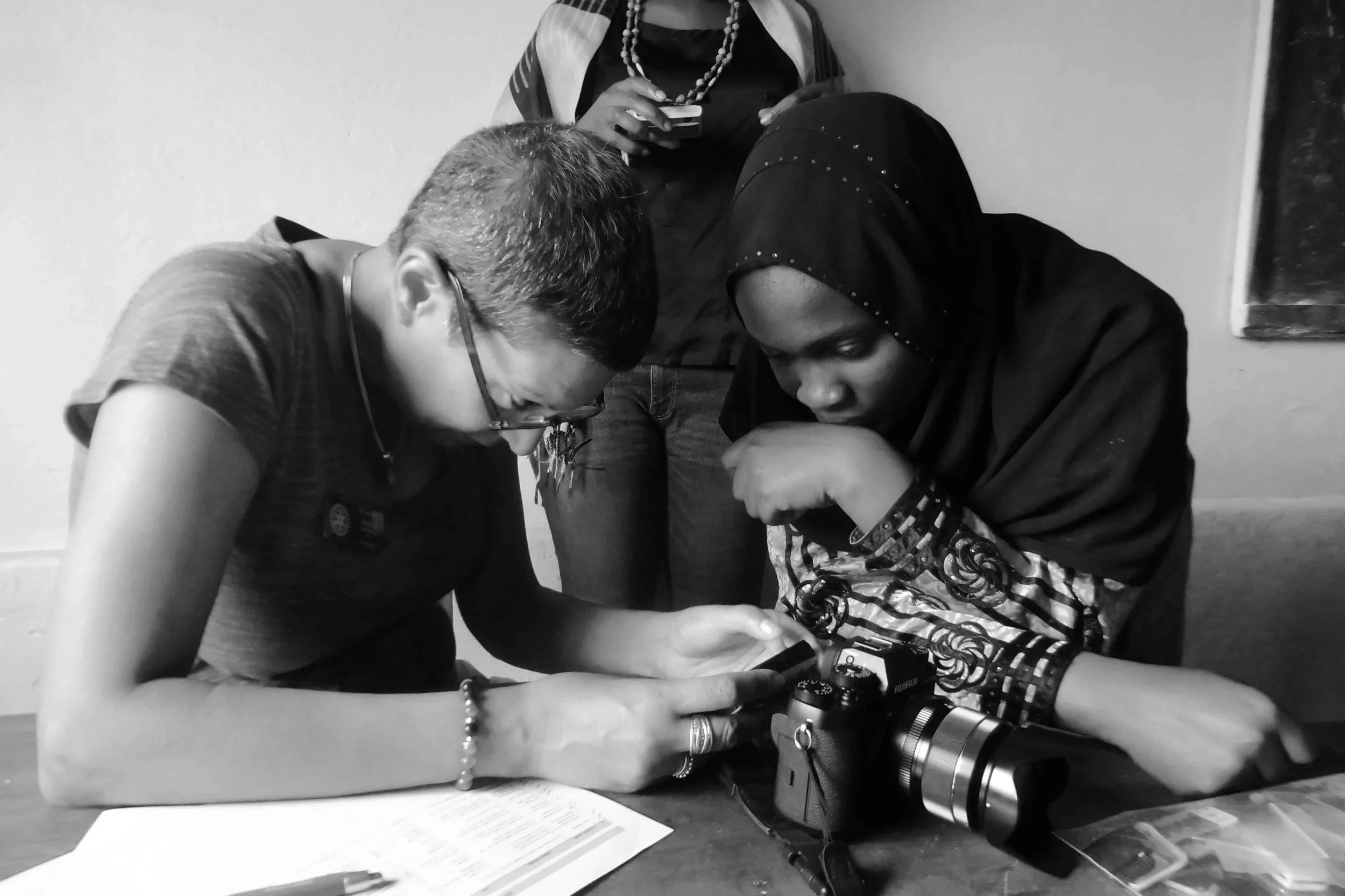 Two women, one with light skin and one with dark skin wearing a headscarf, are leaning over a table looking at a smartphone. There is a camera on the table, papers, and writing utensils.