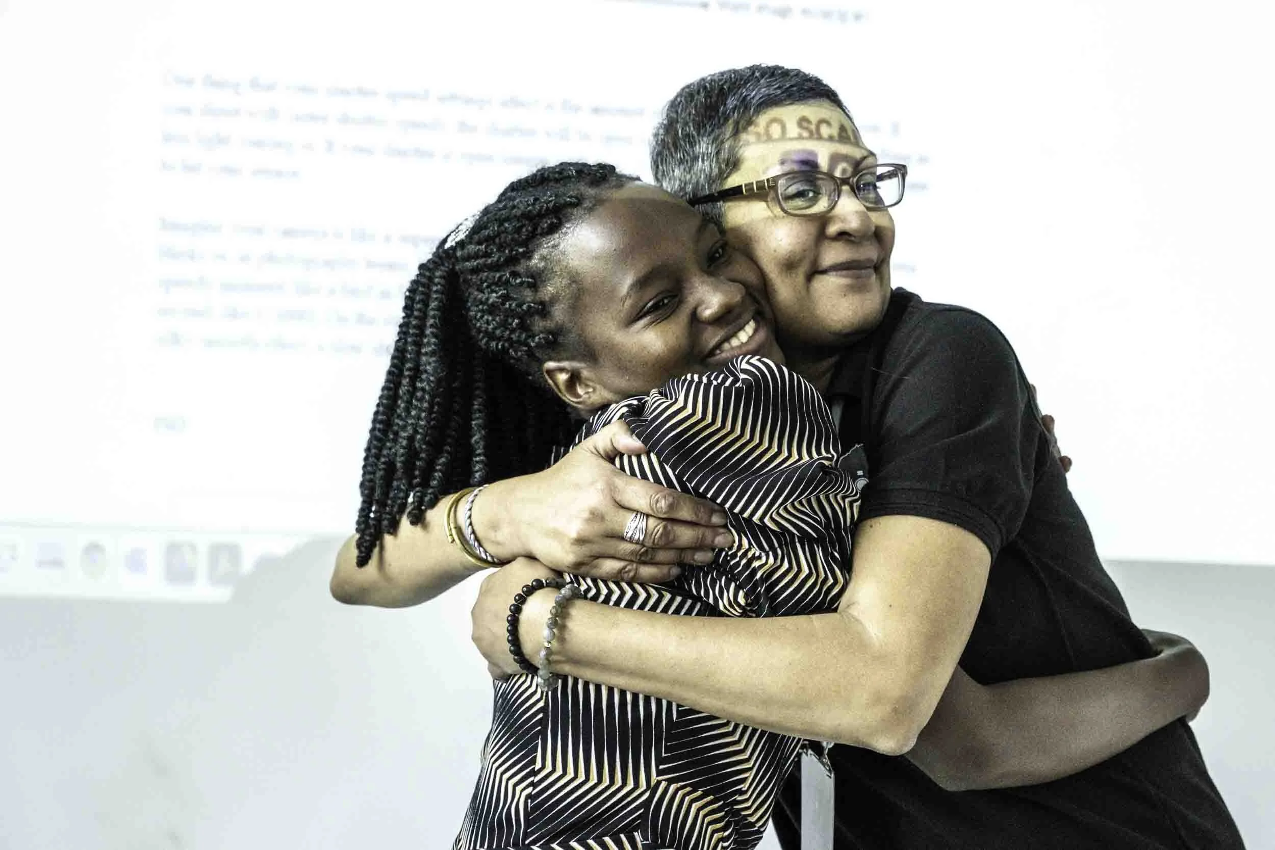Two women embracing and smiling, one with braids and the other with short gray hair and glasses, standing in front of a white background with some text.