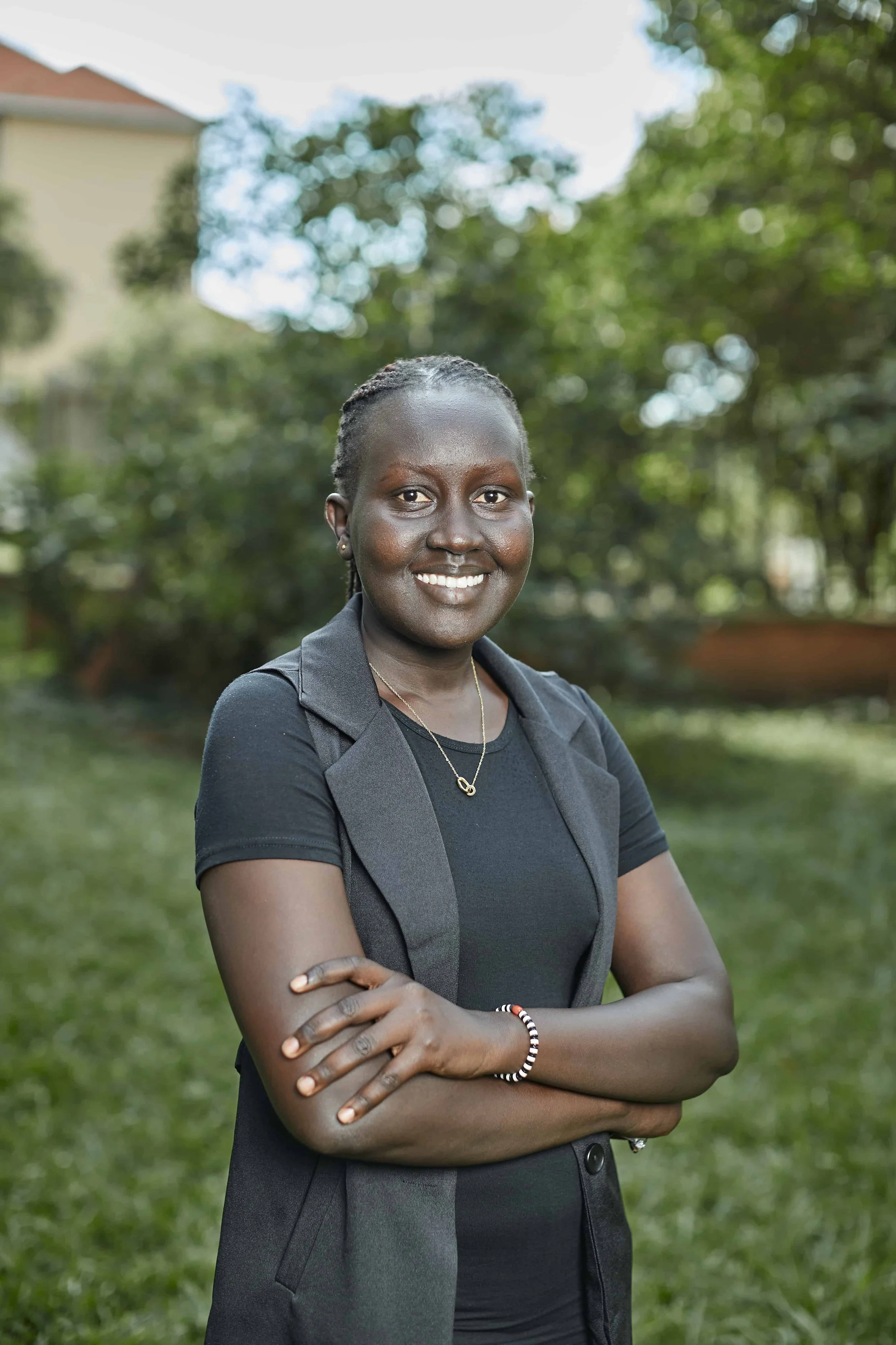 Portrait of a smiling woman with dark skin, short braided hair, wearing a black T-shirt, a dark blazer, and a necklace, standing outdoors with crossed arms in a park with green trees and a building in the background.