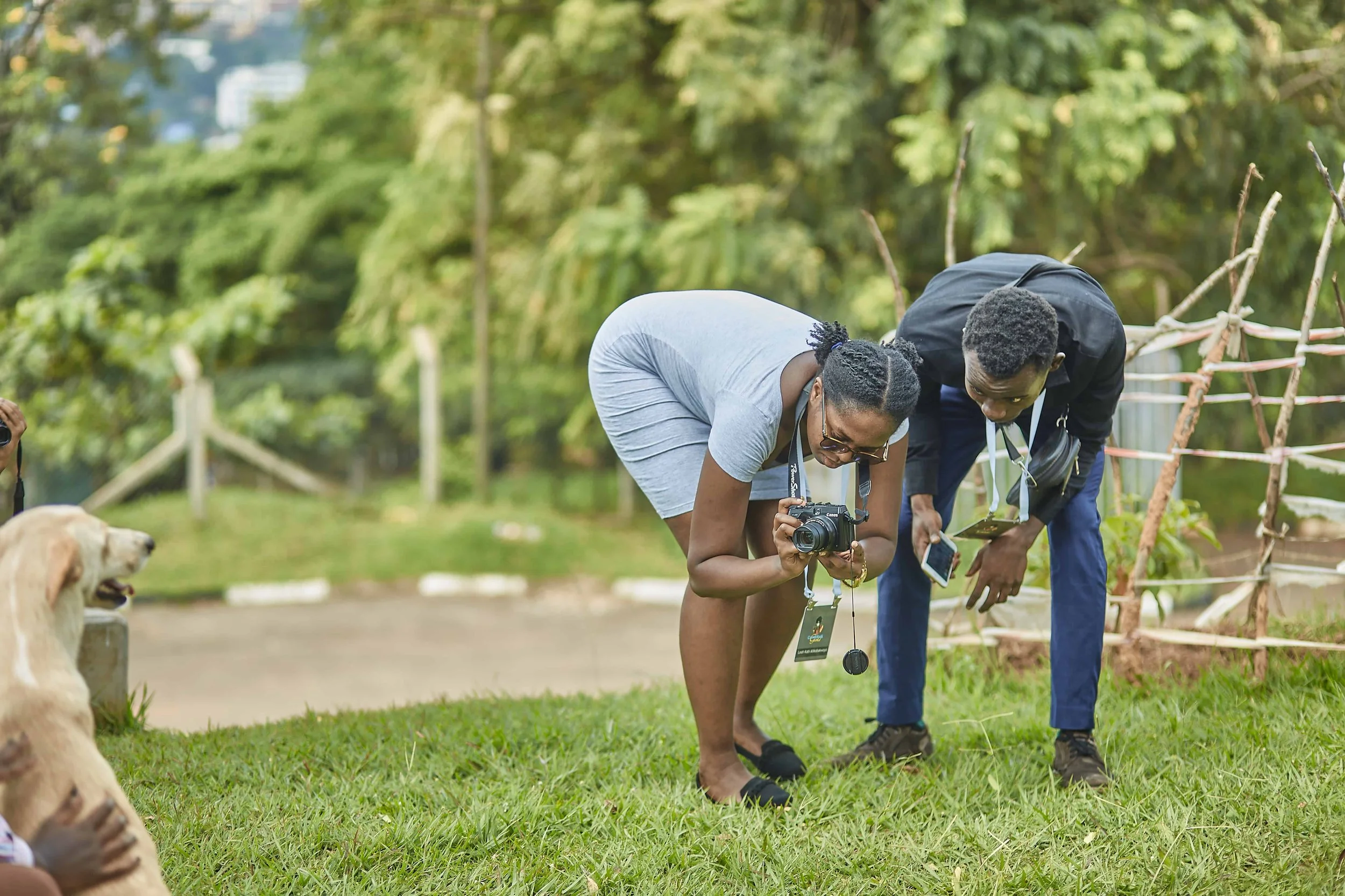 Two women are examining objects on the ground outdoors in a green park. One woman is holding a camera, wearing glasses and a gray shirt, and the other woman is wearing a black jacket and pants. A dog is visible in the foreground, and trees and a path