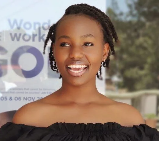 A woman with short braided hair smiling outdoors, with a blurred background that includes a poster.