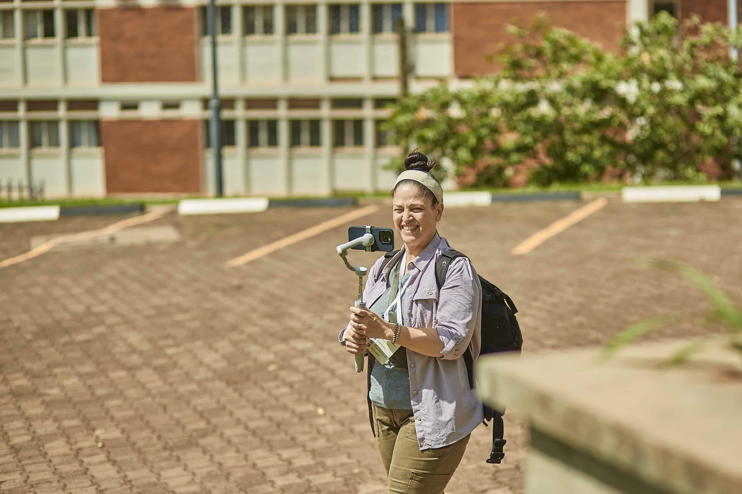 A person smiling and taking a selfie outdoors with a smartphone mounted on a selfie stick. The person is wearing a headband, a grey jacket, and khaki pants, and is carrying a backpack. The background shows a parking lot, green trees, and a building.