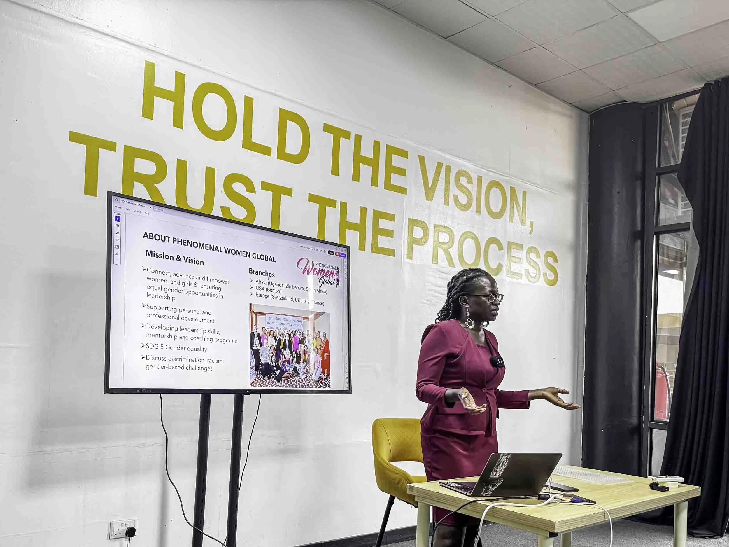 A woman in a pink blazer giving a presentation or speech in an indoor setting with a large screen displaying information about Phenomenal Women Global. The background includes a wall with a quote in large yellow letters saying "HOLD THE VISION, TRUST