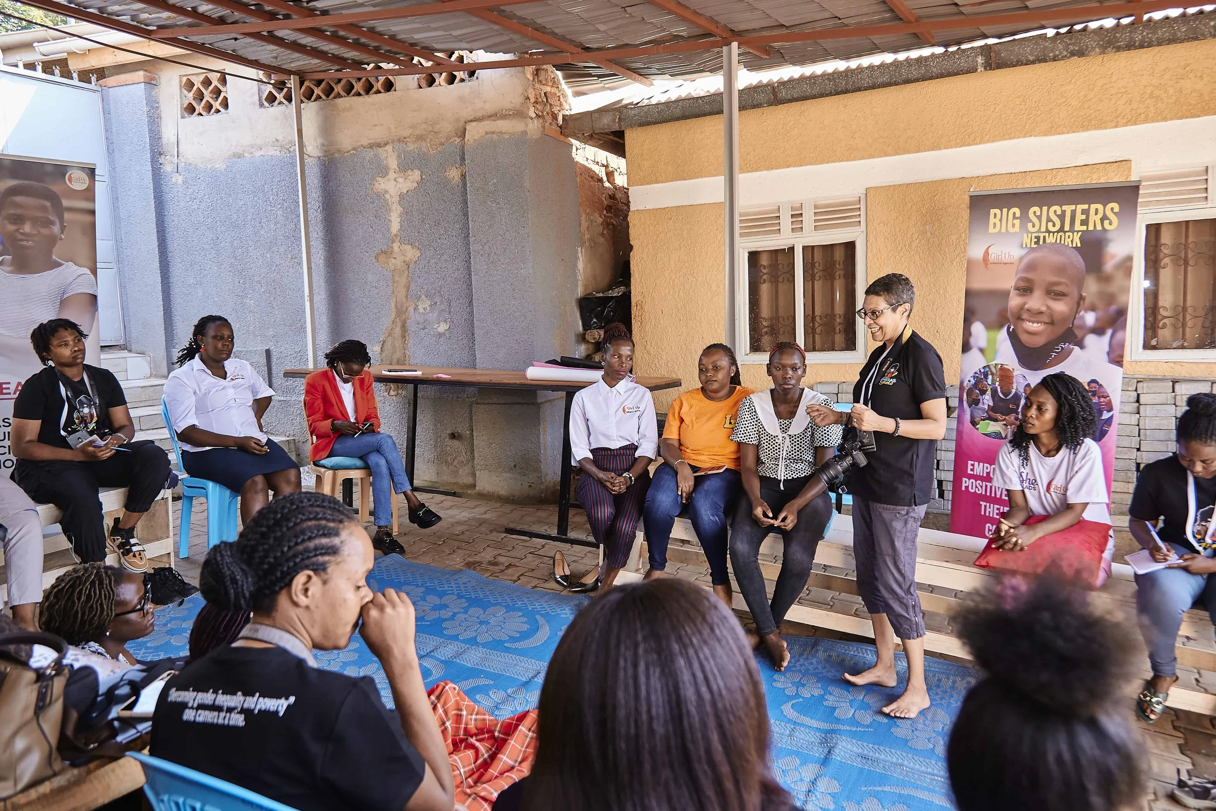 A group of women and girls seated outdoors during a community event, with some standing and speaking, banners in the background, and a partially unfinished wall behind them.