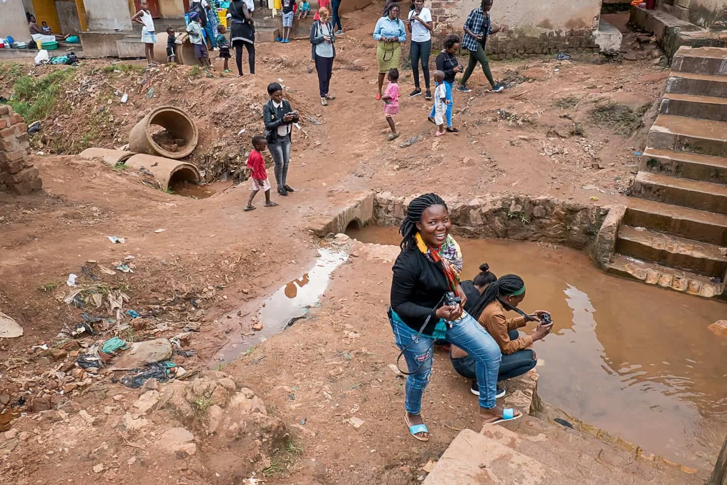 A woman smiling and holding a camera standing near a small water pool, with two women photographing the water, children around, and structures under construction or ruin in a dirt area.