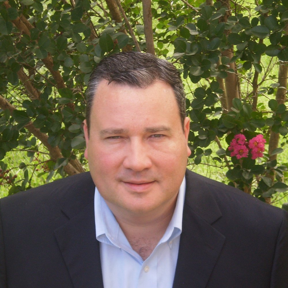 A man in a black suit jacket and white shirt is outdoors, surrounded by green leaves and pink flowers, looking at the camera.