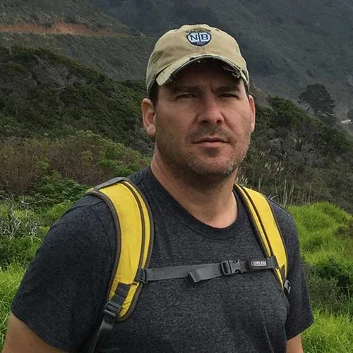 Man in a black t-shirt with a yellow backpack, wearing a beige cap, standing outdoors in a hilly, green landscape.