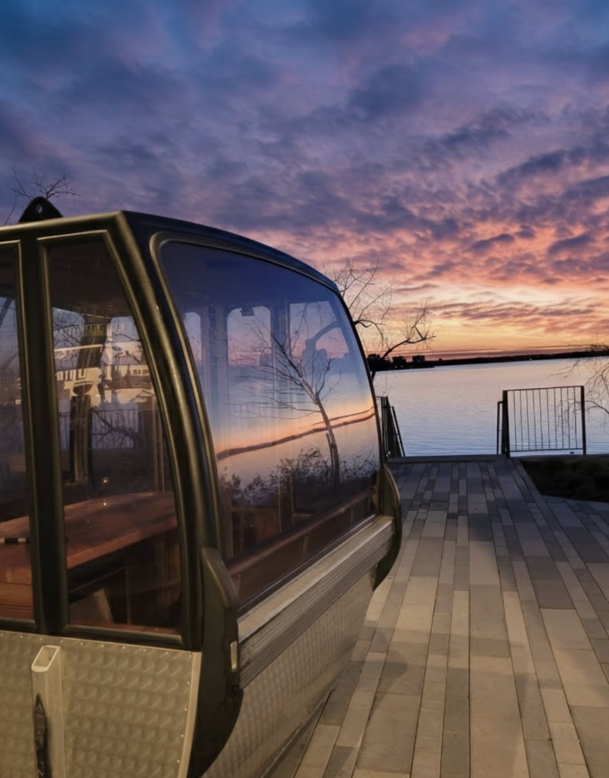 A gondola lift parked on a waterfront promenade at sunset, with colorful clouds and waters reflecting the sky in the background.
