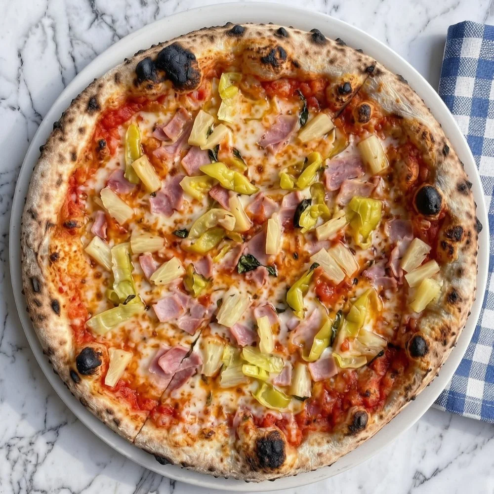 A freshly baked pizza with cheese, ham, artichoke hearts, chicken and green peppers on a white plate, placed on a marble surface with a blue and white checkered napkin nearby.