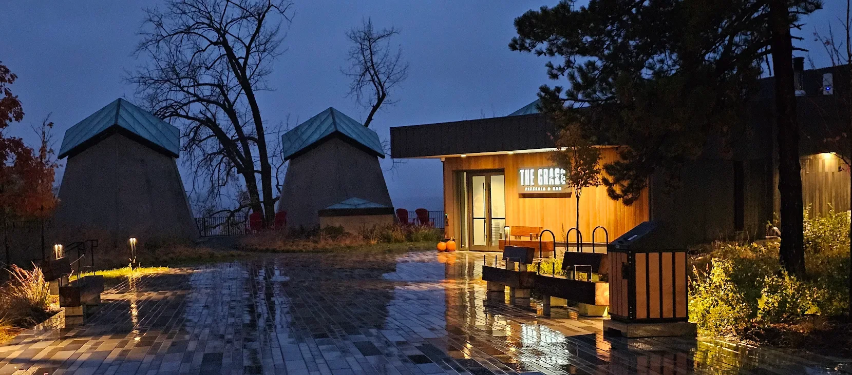 A wet brick patio at dusk with pumpkins and benches outside a building with a sign that reads "The Green," in a park-like setting with trees and two small buildings with conical roofs in the background.