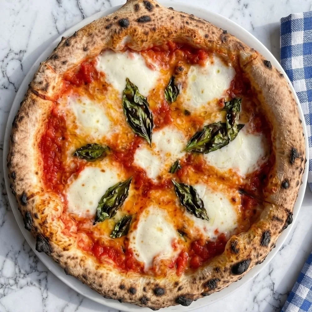 A Margherita pizza topped with fresh basil leaves, mozzarella cheese, and tomato sauce. The pizza has a slightly charred / leoparding on the crust and is served on a white plate on a marble surface at The Grand Pizzeria