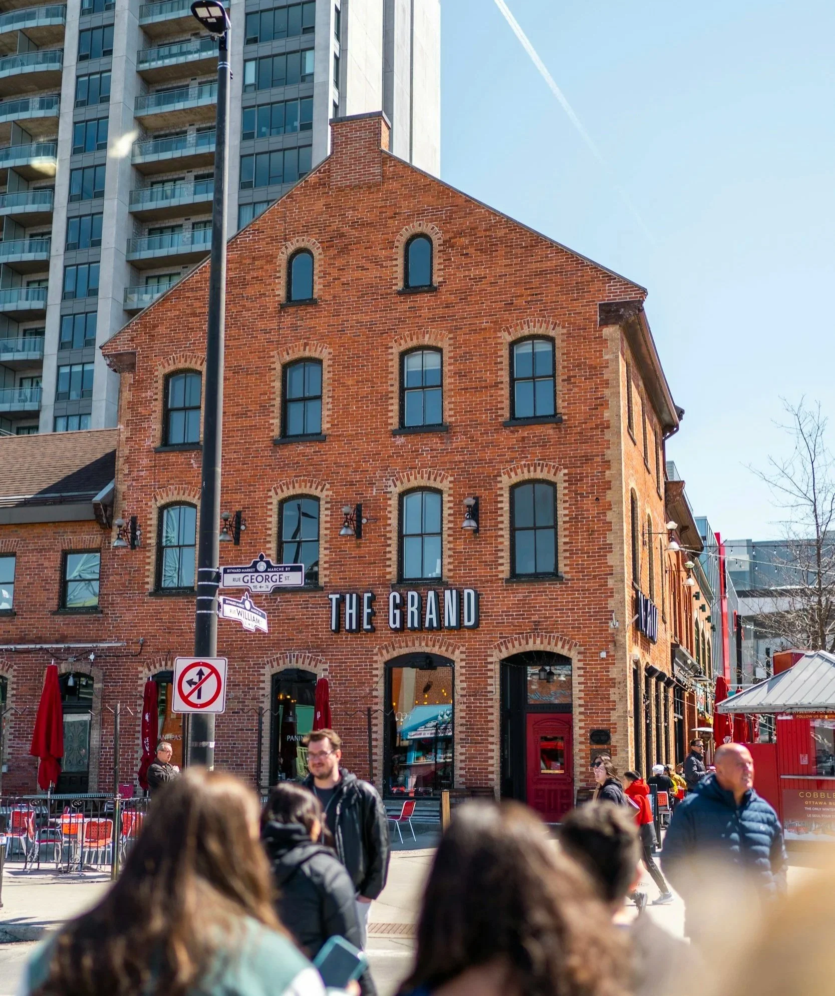 A busy street scene in front of a brick building named 'The Grand', with people walking and sitting outside, and a food truck on the right.